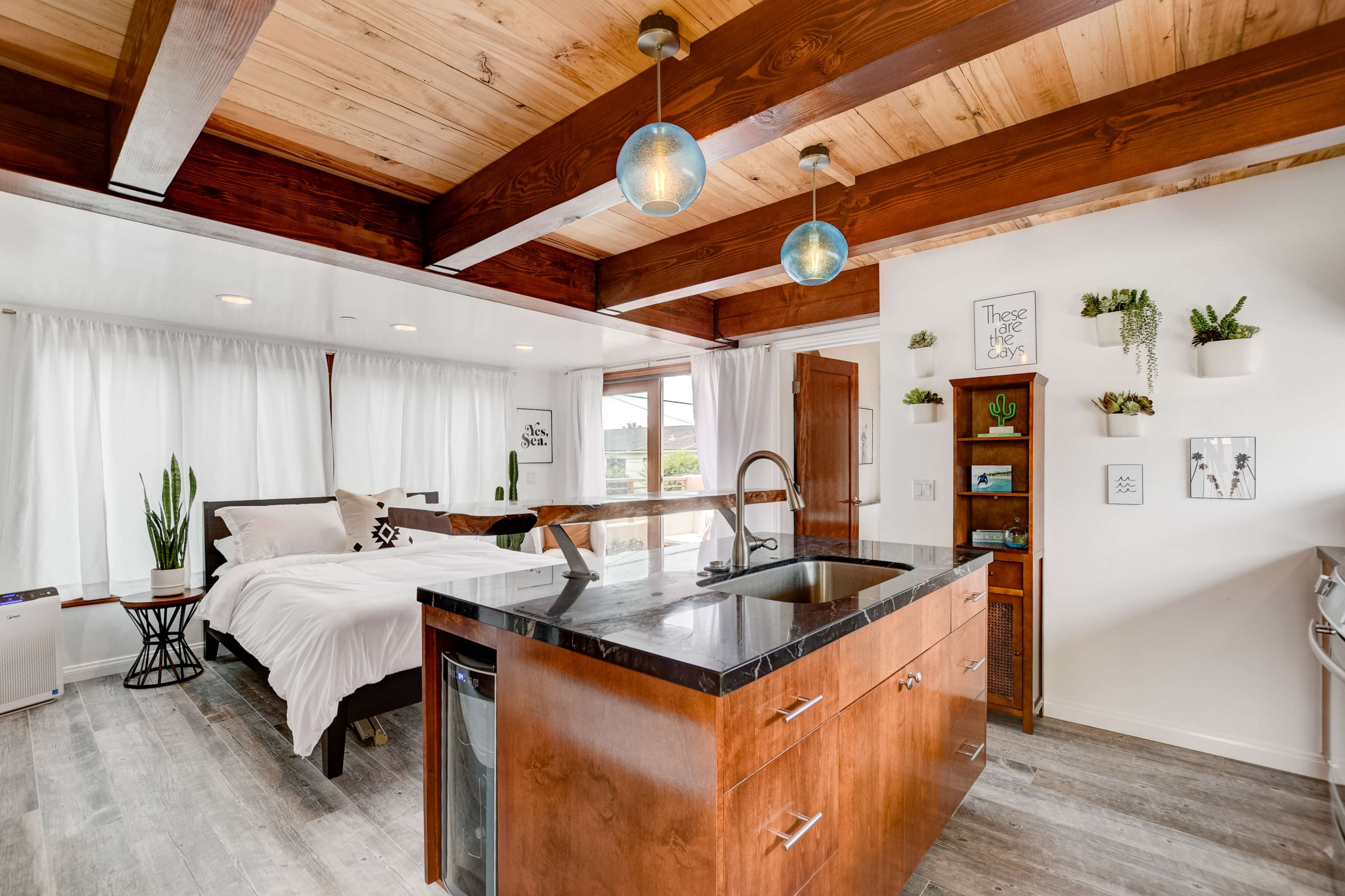 A modern studio apartment with wooden beams, a kitchen island, and a bed area separated by sheer curtains.