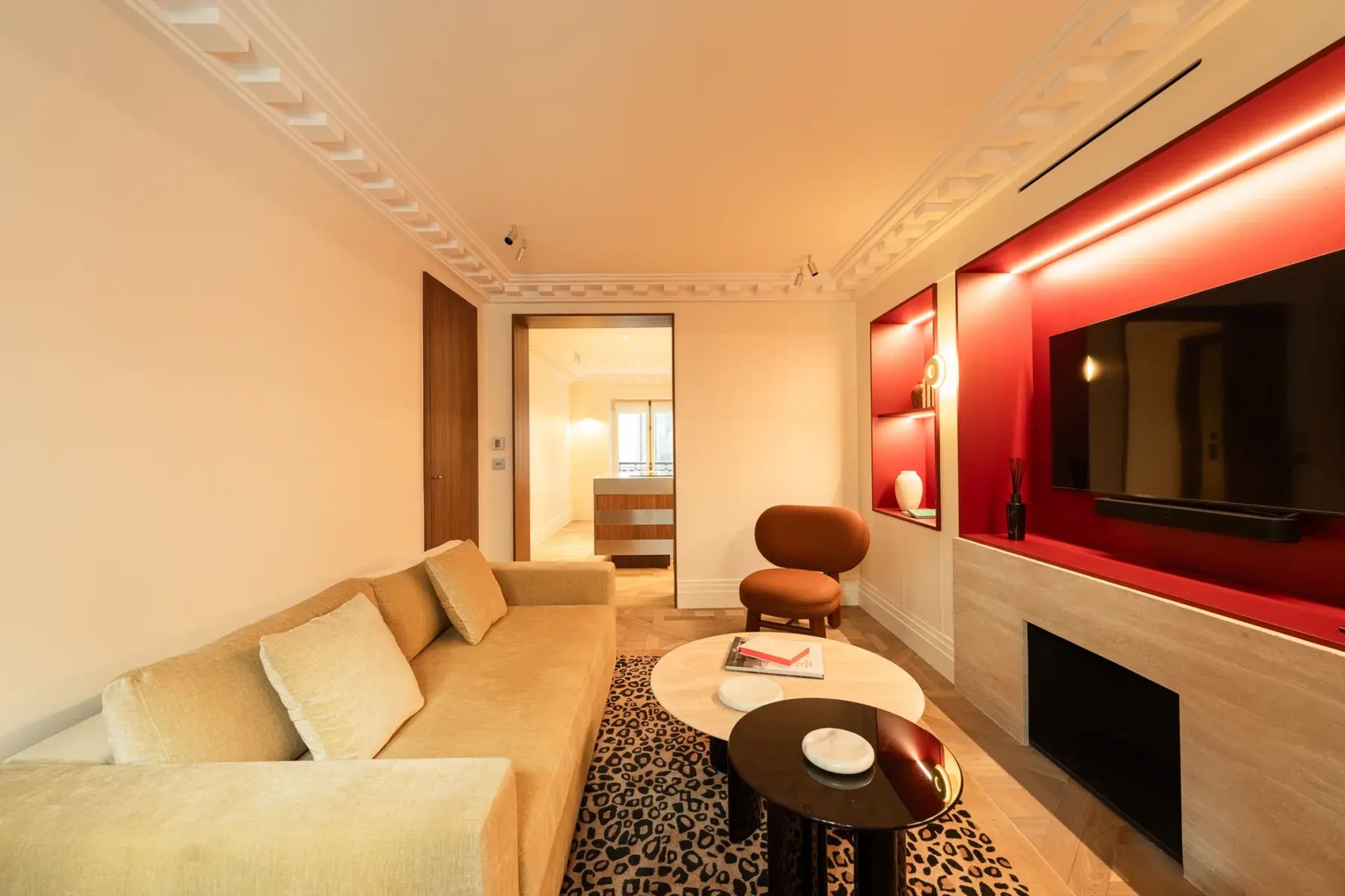 The image shows a modern living room with a beige sofa, a coffee table, a decorative rug, and a flat-screen TV mounted on a wall with red accents.