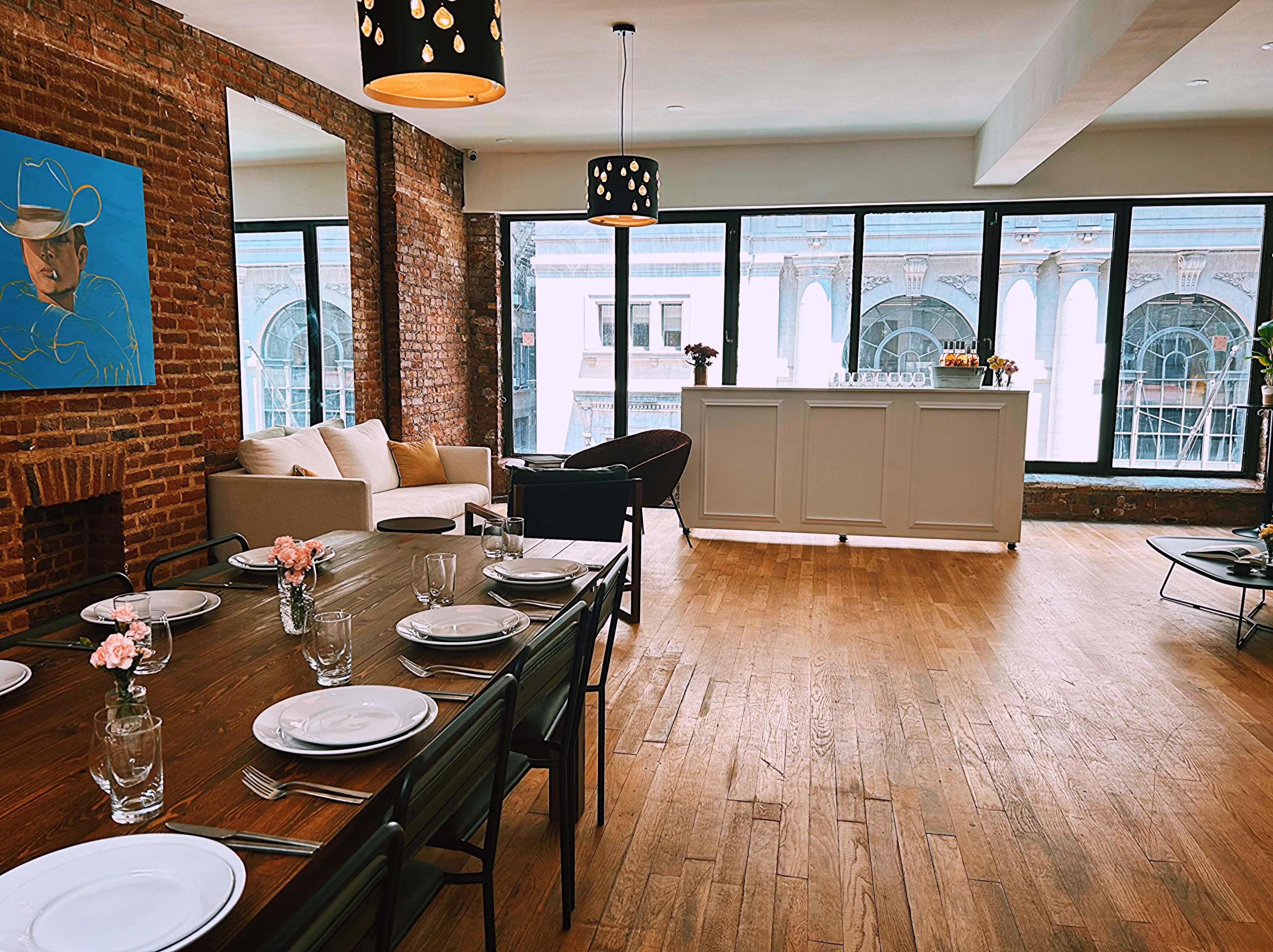 A spacious, well-lit room with a wooden dining table set for a meal, a bar area at one end, and large windows overlooking a city street.