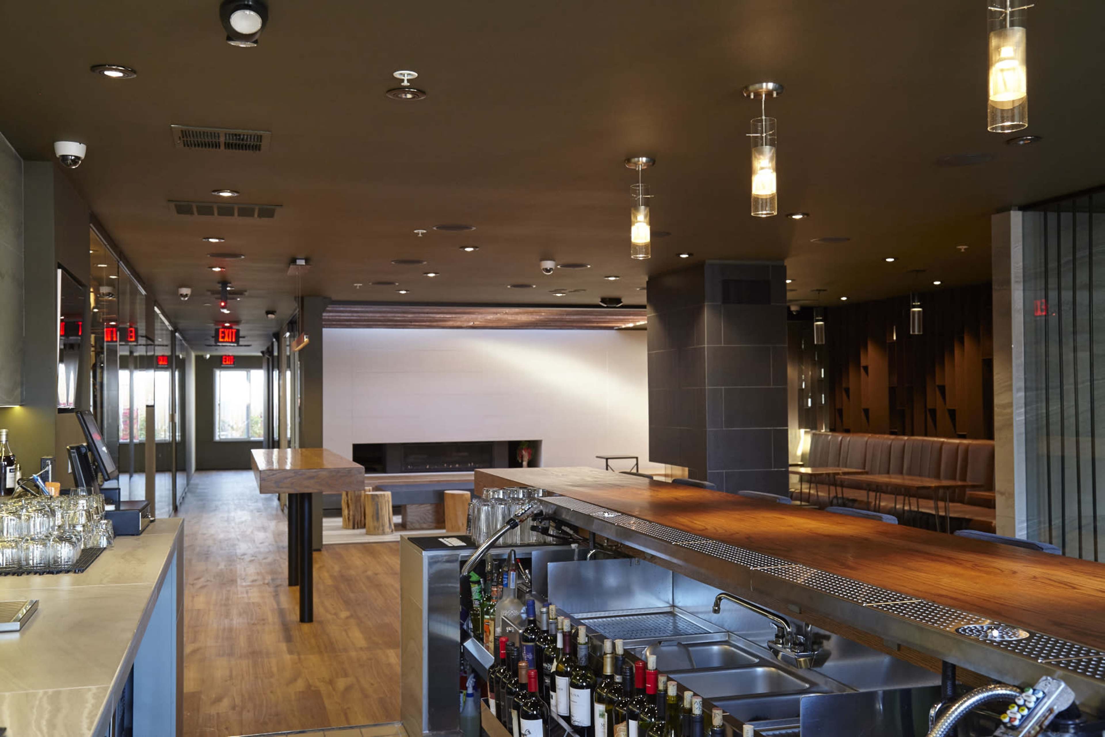 The image shows a modern bar area with a wooden countertop, metallic bar equipment, and a spacious seating area featuring a fireplace and contemporary design elements.