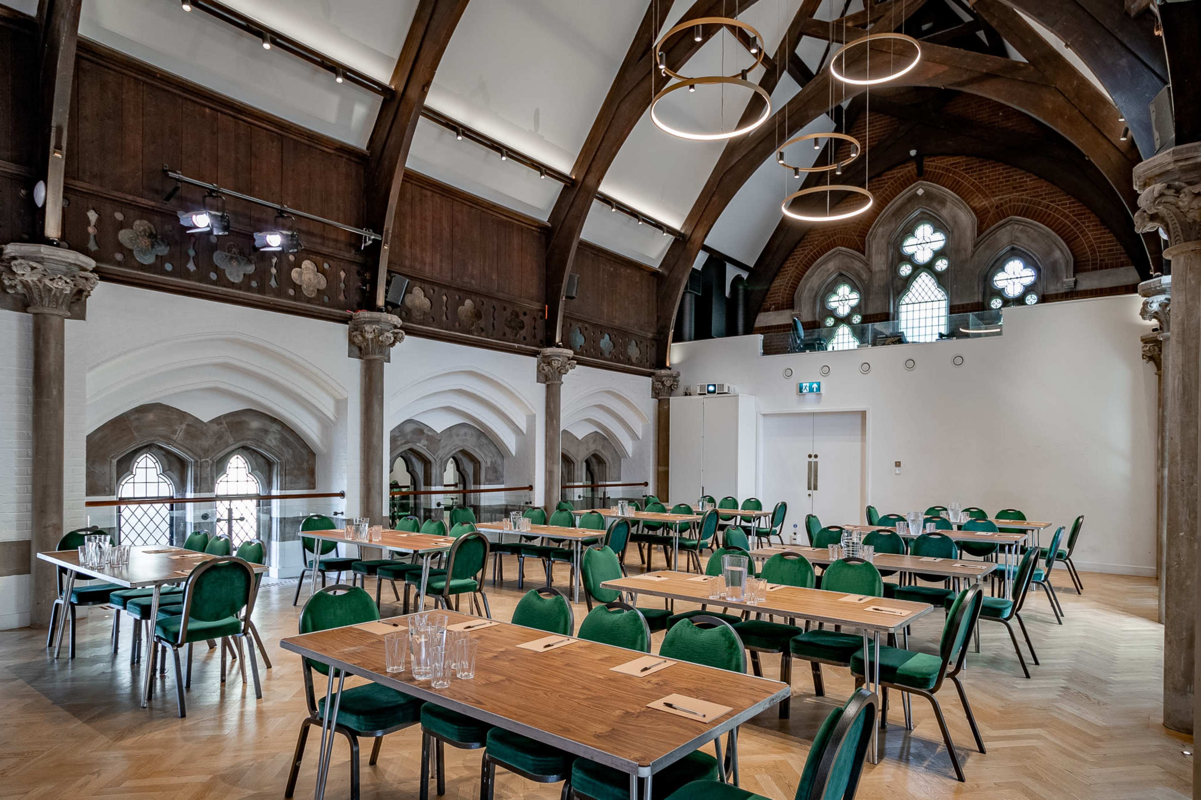 The image shows a spacious dining hall with wooden beams, multiple tables set with glassware, and large arched windows.