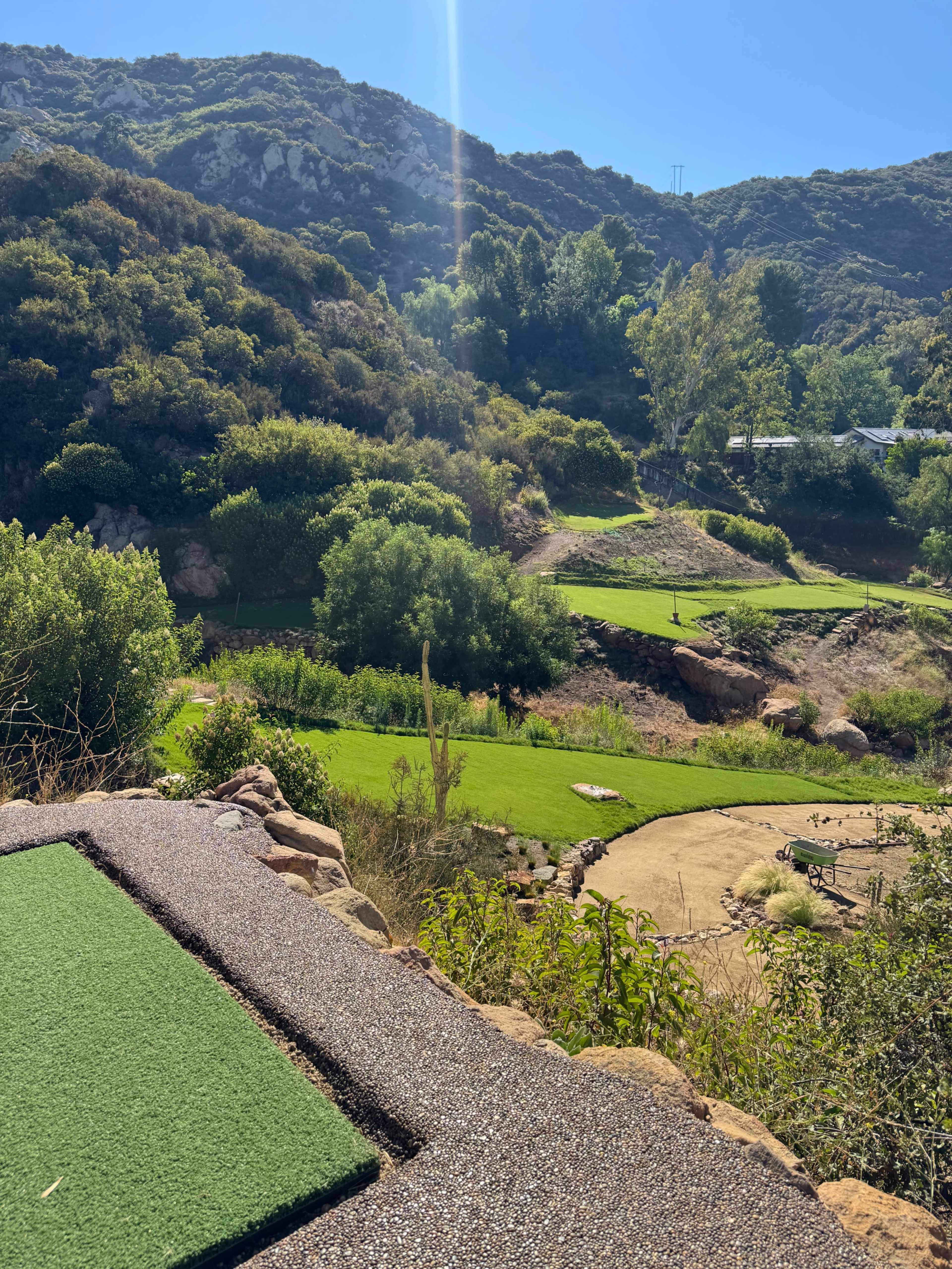 The image shows a landscaped area with several green plots of grass situated among rolling hills under a clear blue sky.