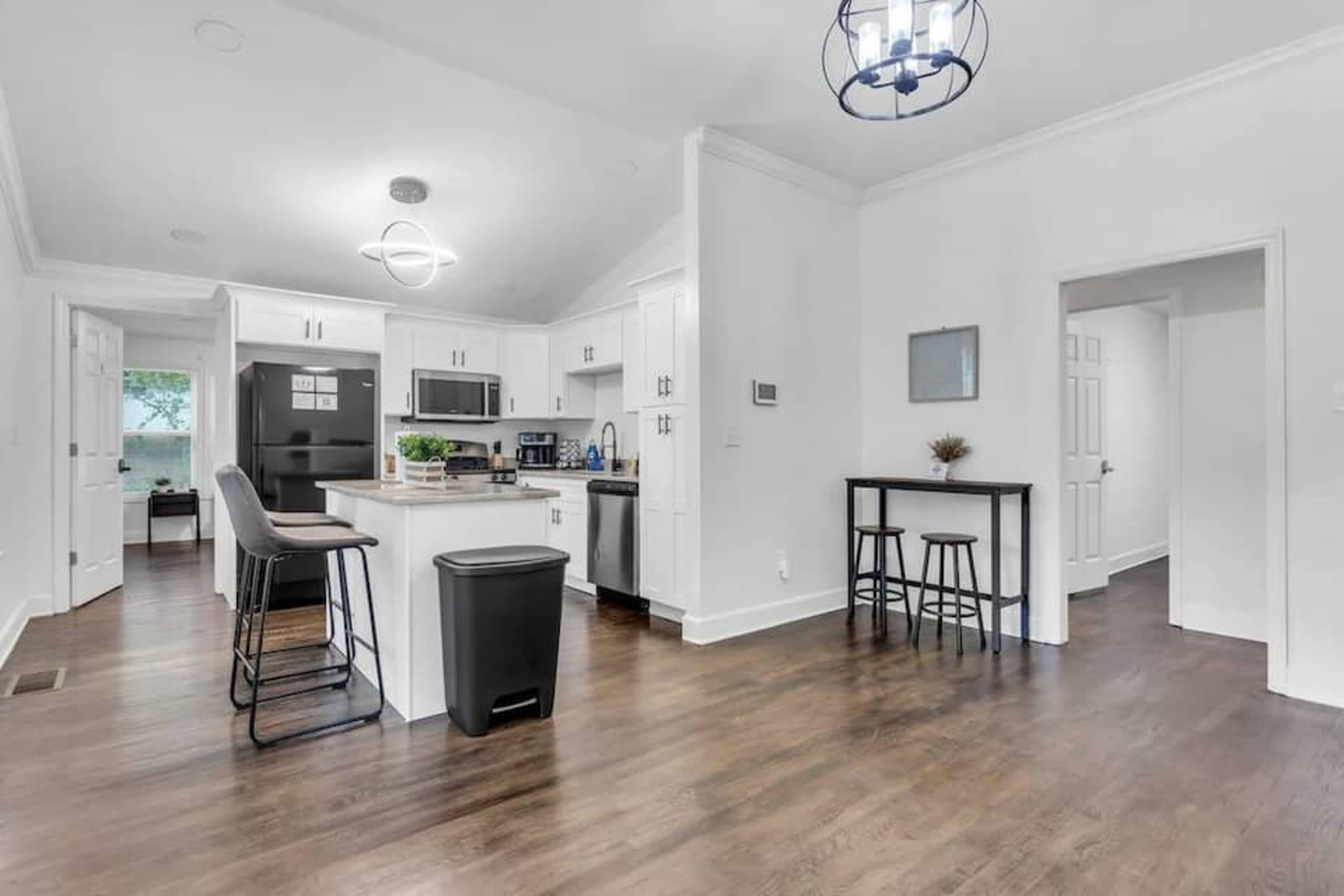 A modern kitchen area with white cabinetry, stainless steel appliances, a breakfast bar with stools, and a small dining table in an open layout.