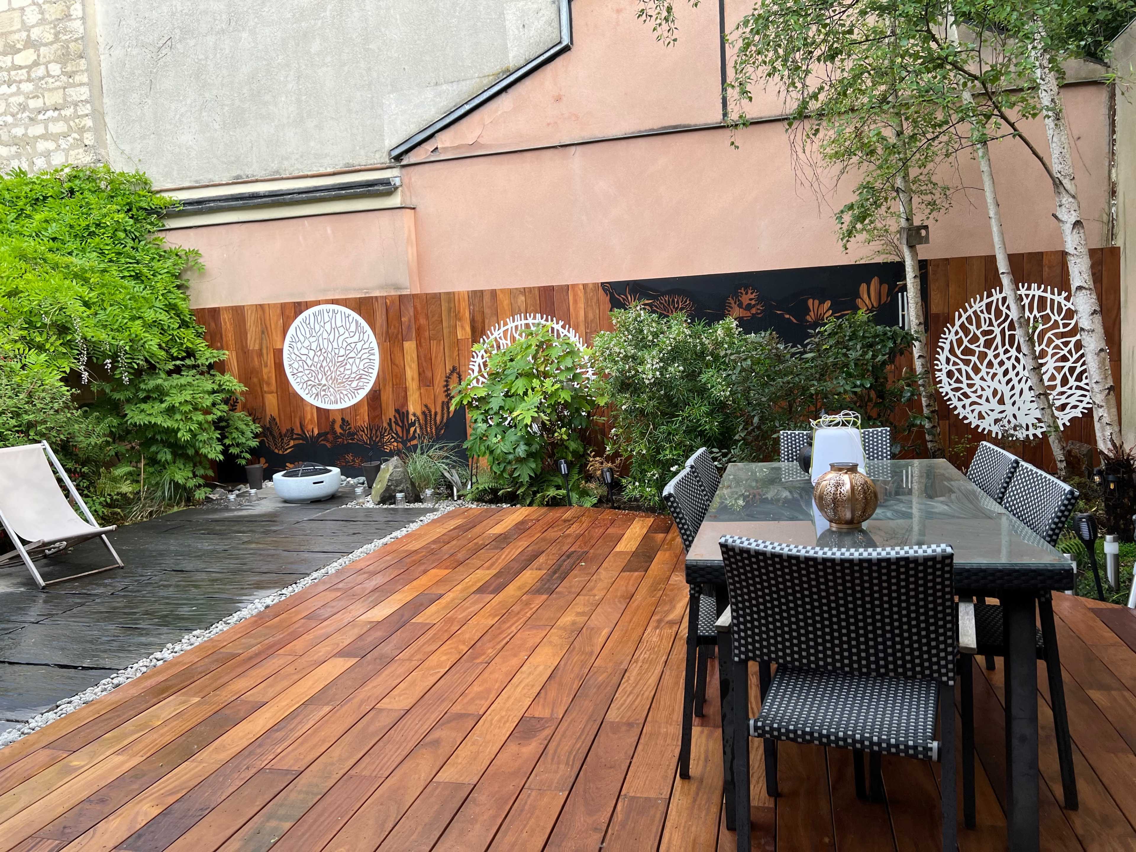 A wooden deck with a glass dining table surrounded by black wicker chairs, set against a backdrop of greenery and patterned wall art.
