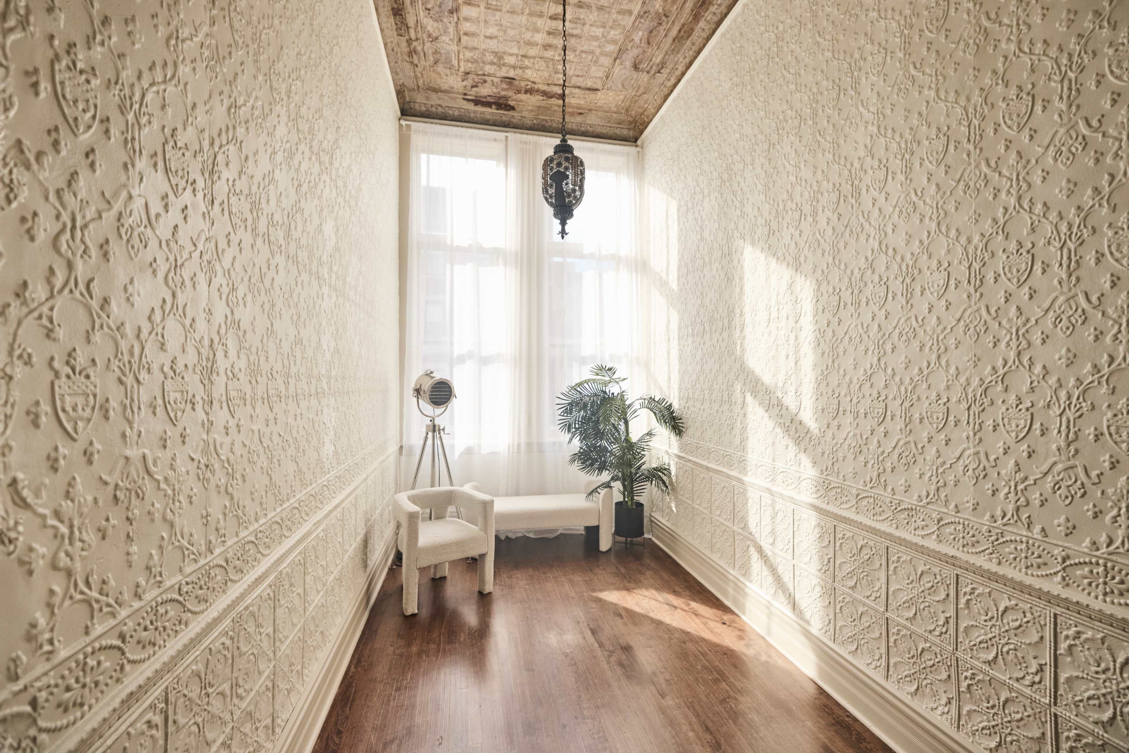 A narrow hallway with patterned white walls, a wooden floor, a chair, a potted plant, and a large window allowing natural light to enter.
