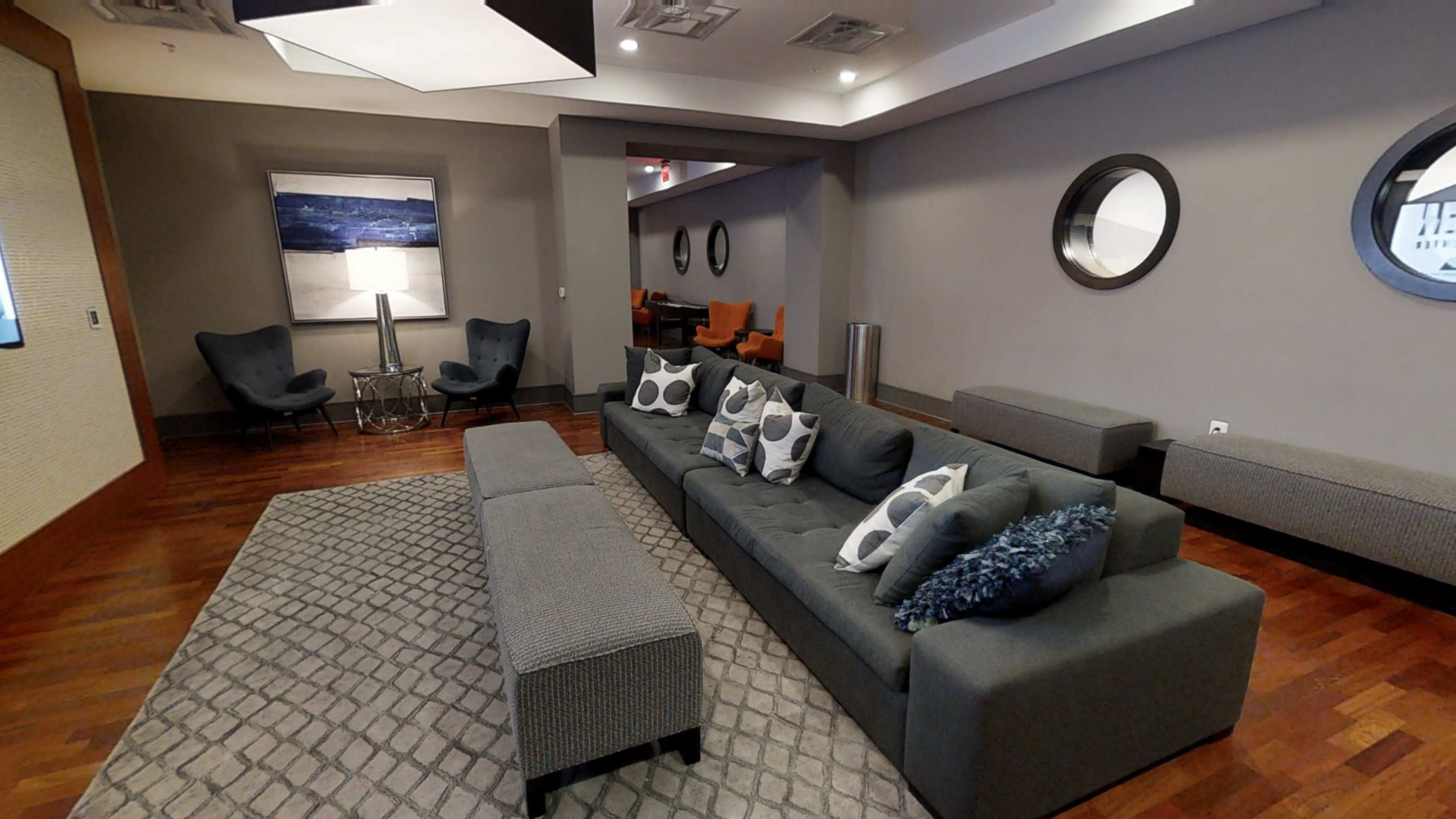 A modern living area with a gray sectional sofa, a patterned rug, and various seating arrangements, including chairs and tables against a neutral wall.