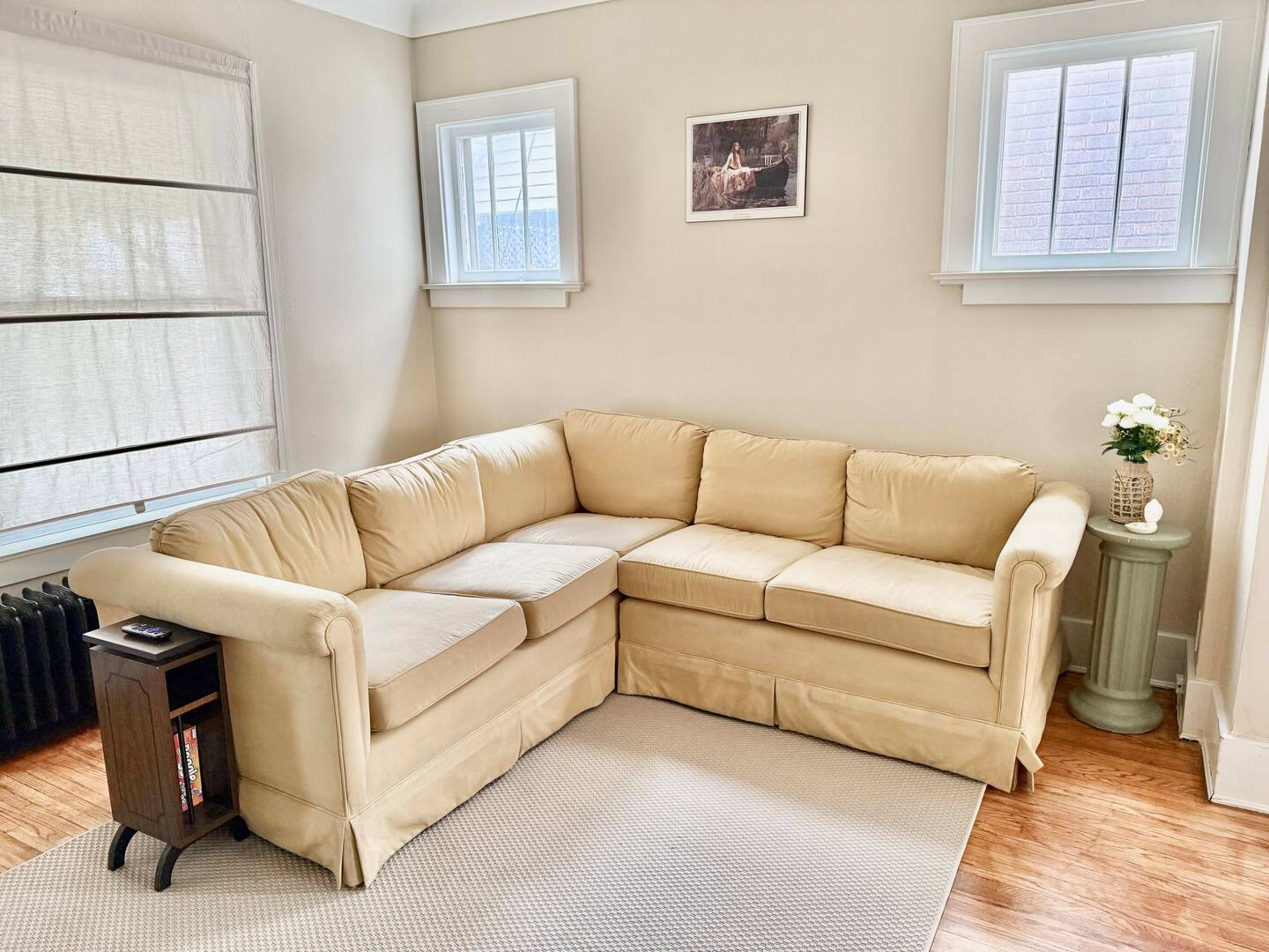 The image shows a light-colored sectional sofa positioned in a well-lit living room with two windows and a small table beside it.