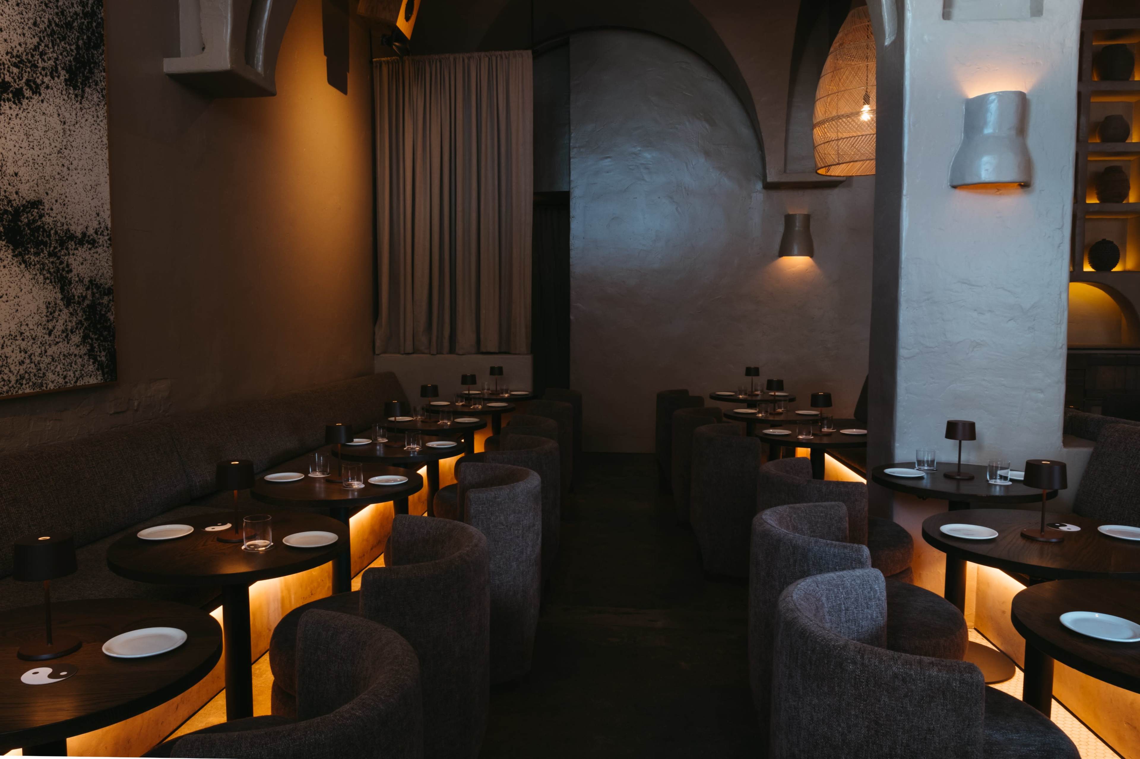 A dimly lit restaurant features several circular tables surrounded by plush, gray chairs against a backdrop of textured walls and softly glowing lamps.