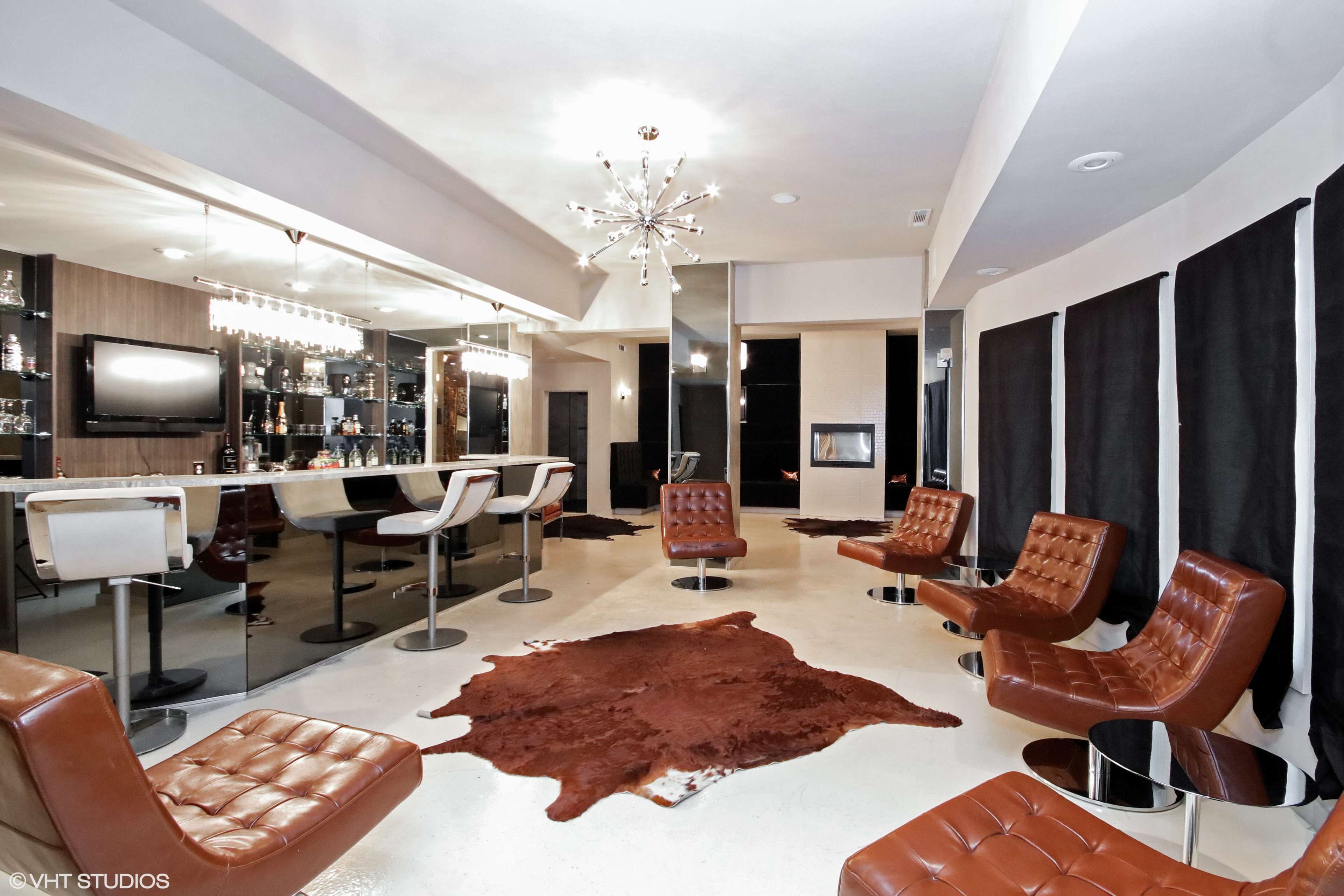 A modern lounge area features a bar with stools, leather seating, and a decorative chandelier against a backdrop of dark window coverings.