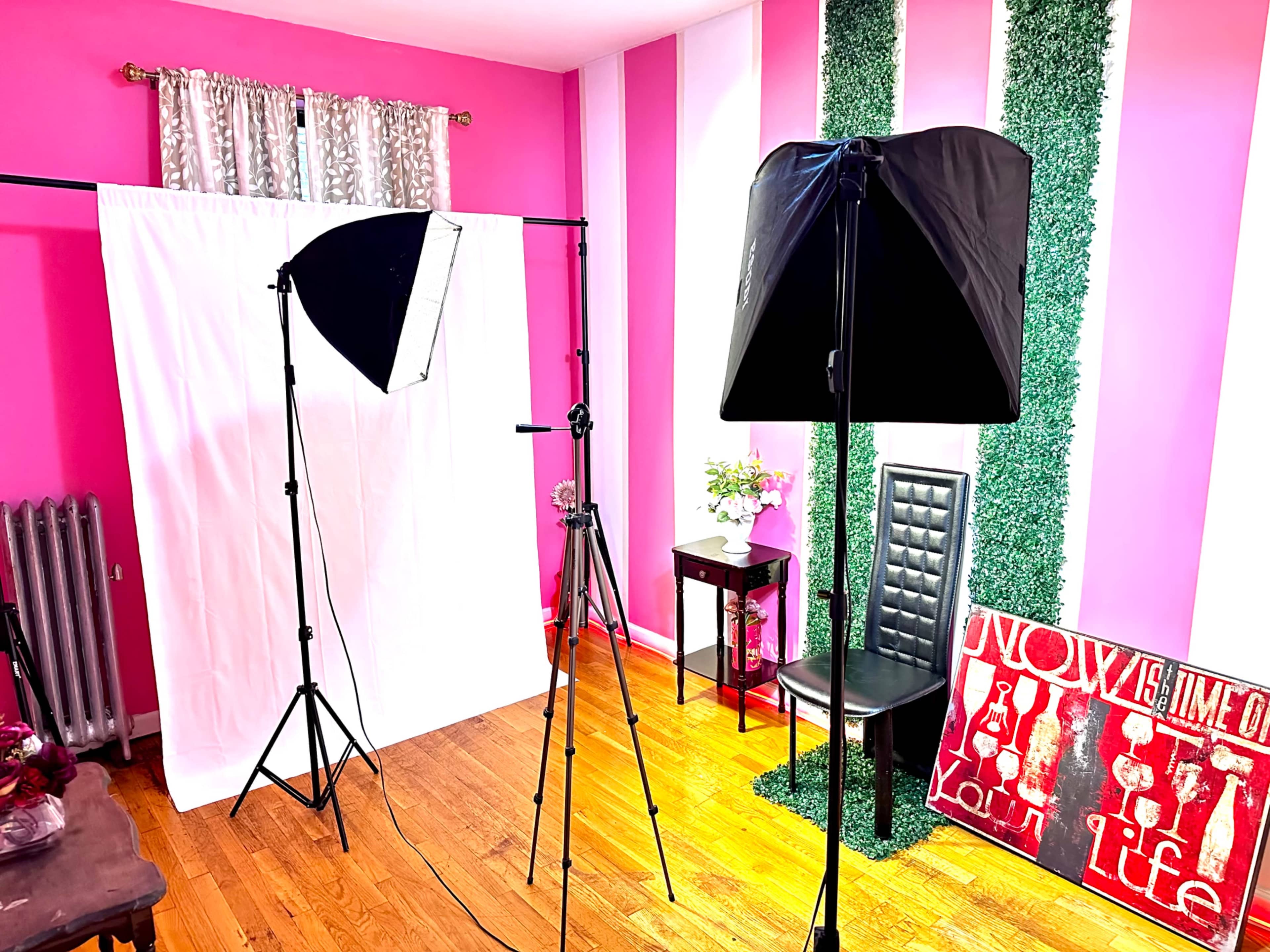 The image features a brightly colored studio setup with a white backdrop, lighting equipment on stands, a black chair, and decorative elements on the walls.