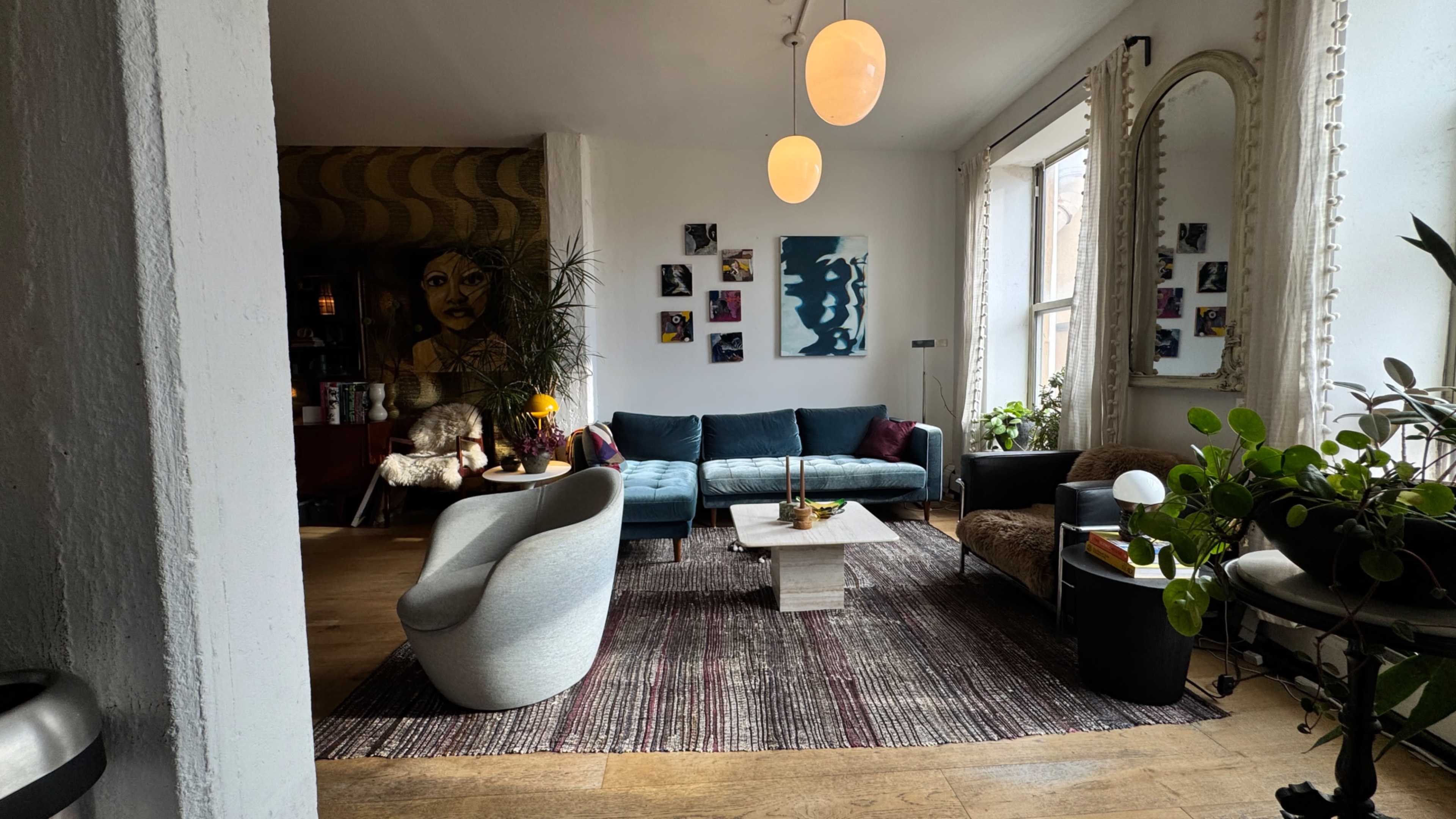The image shows a modern living room with a teal sofa, a light-colored coffee table, and various plants, framed artwork, and warm lighting.