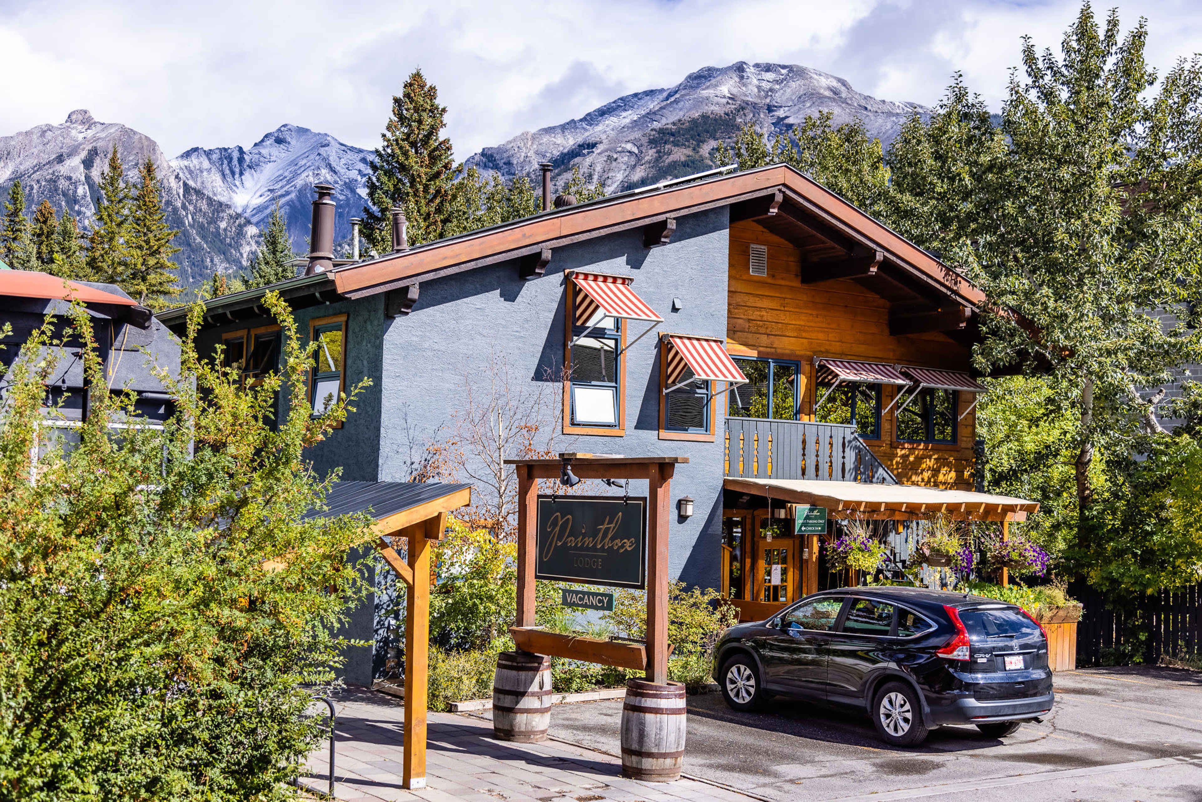 A two-story blue and wooden building with a sign that reads "Vacancy" and a parked car is situated in front of a backdrop of mountains and trees.