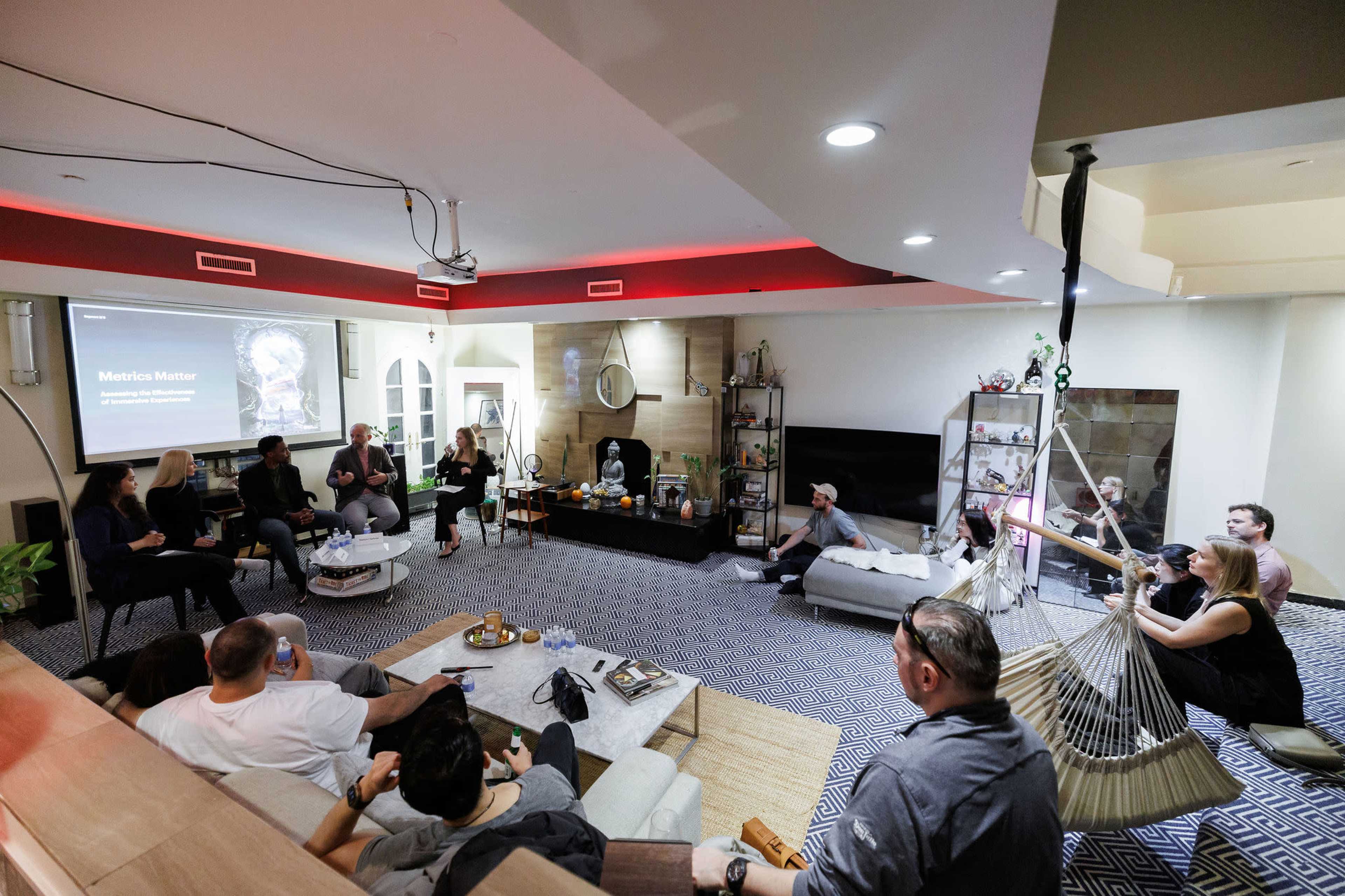 A group of people is seated in a modern living room, engaged in discussion while a presentation is displayed on a screen.
