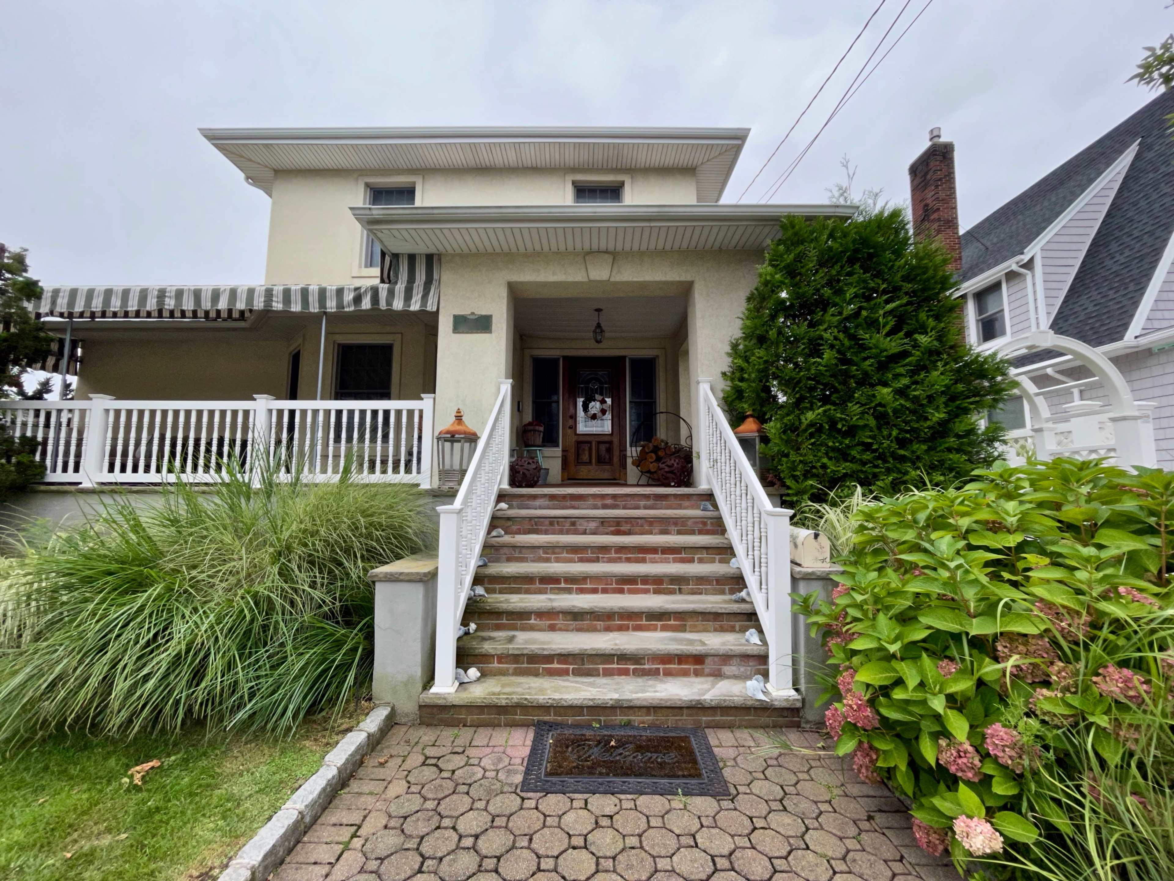 A two-story house with a front porch and steps leading to the entrance, surrounded by shrubs and flowers.
