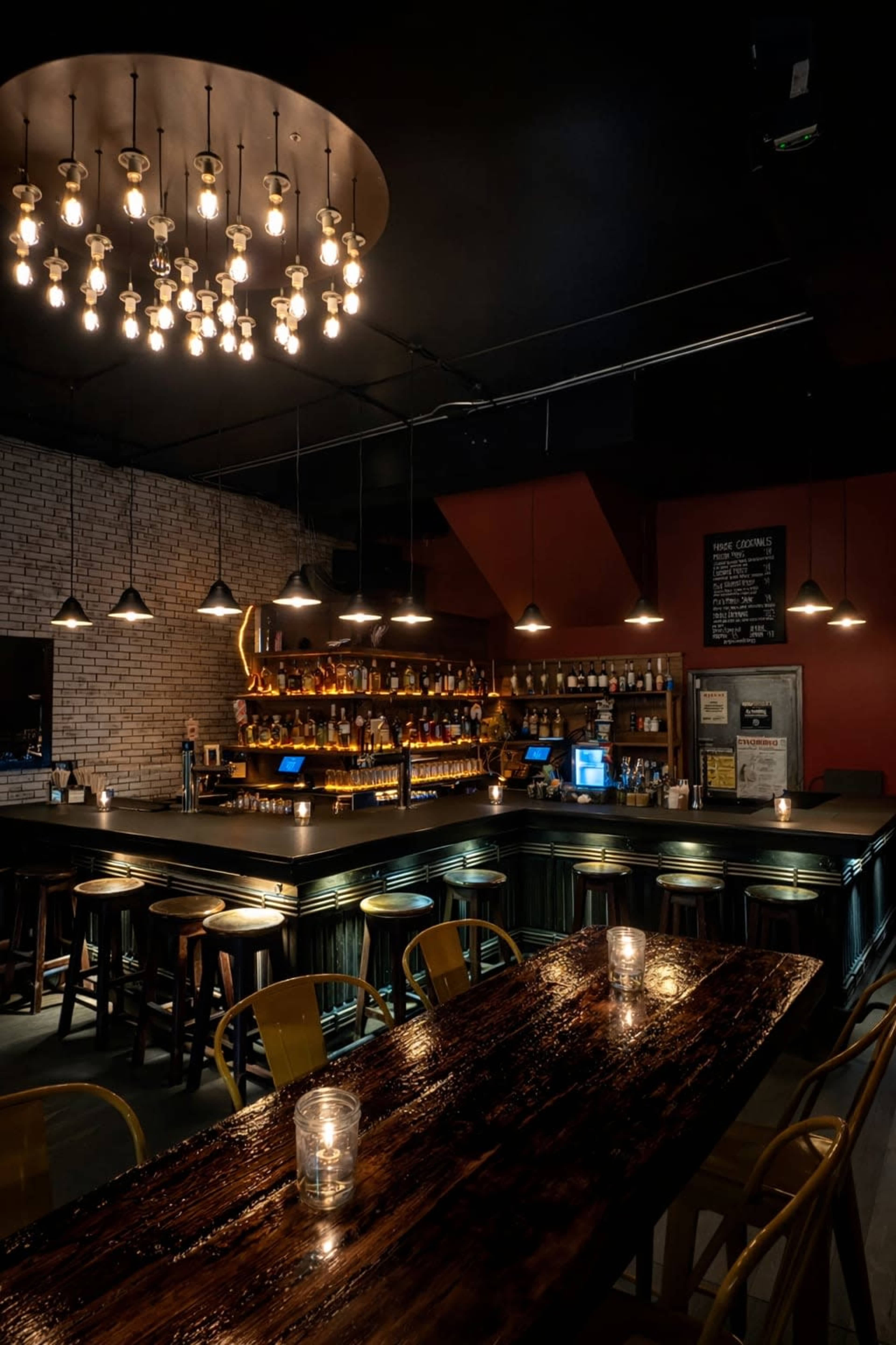 A dimly lit bar features a wooden table with candles, a lengthy counter stacked with various bottles, and an array of pendant lights overhead.