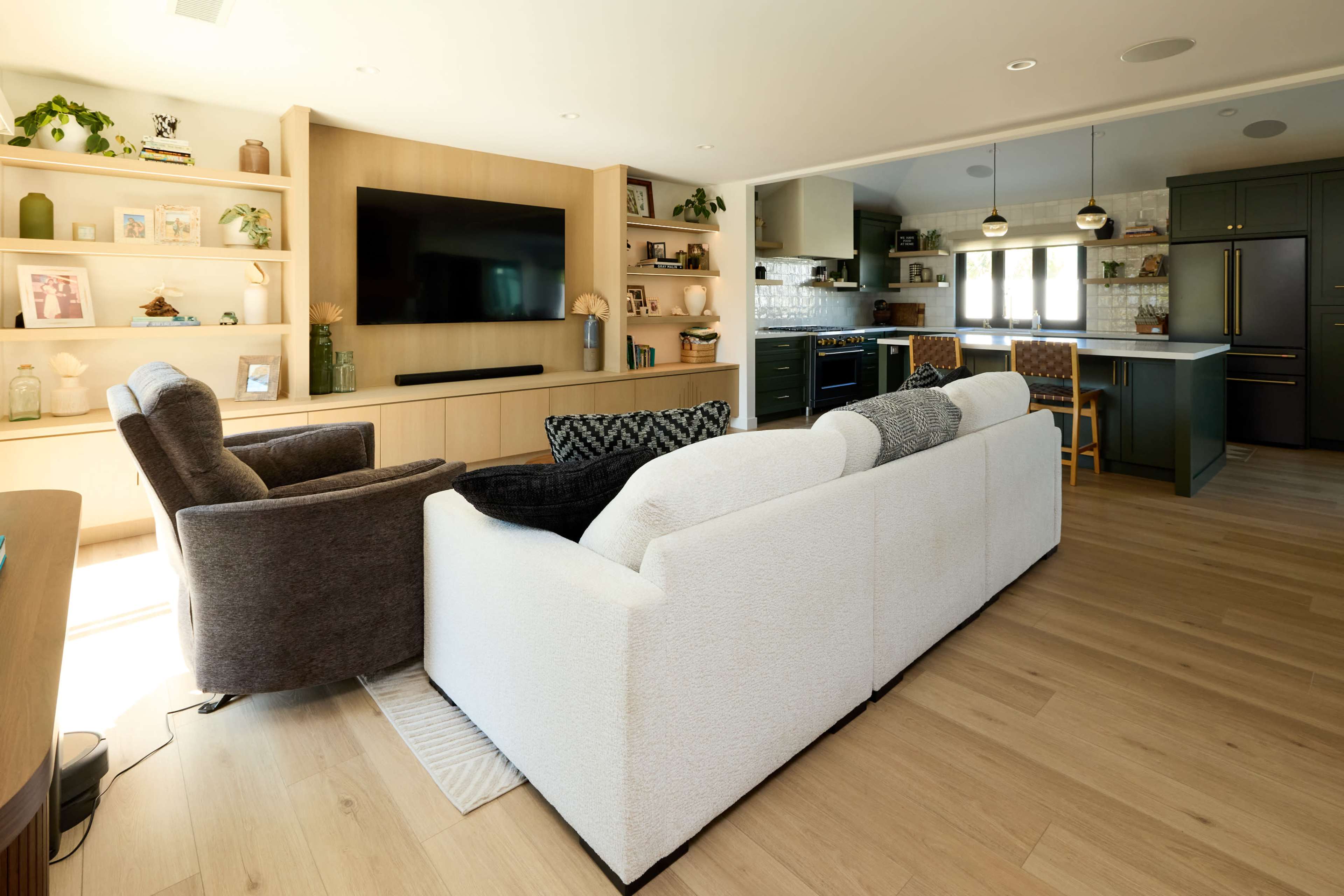 The image shows a modern living room with a white sofa, gray armchairs, a television mounted on a wooden wall, and an open kitchen area in the background.