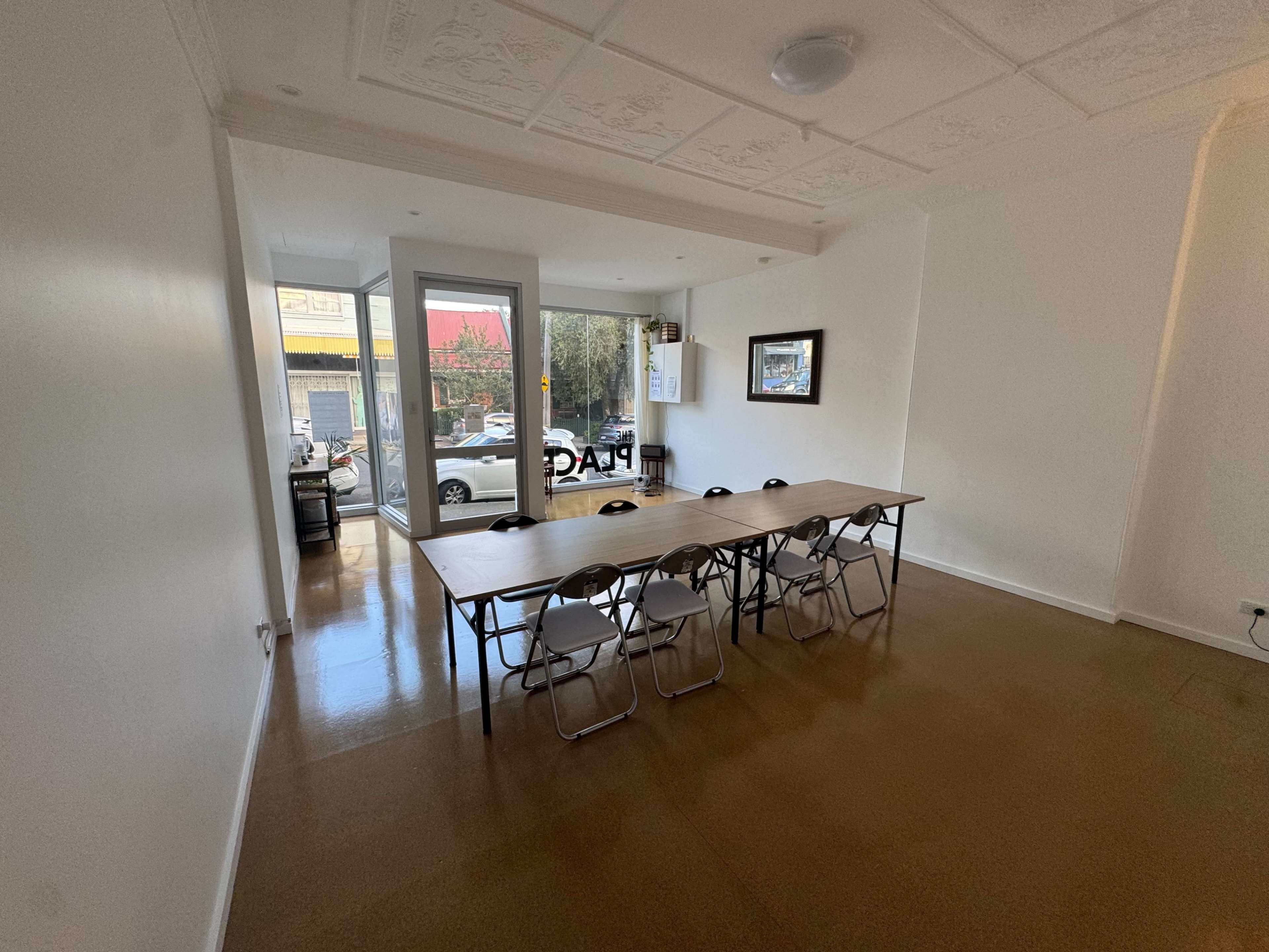 The image shows a modest meeting room featuring a long wooden table surrounded by folding chairs, with a door leading to a street view.