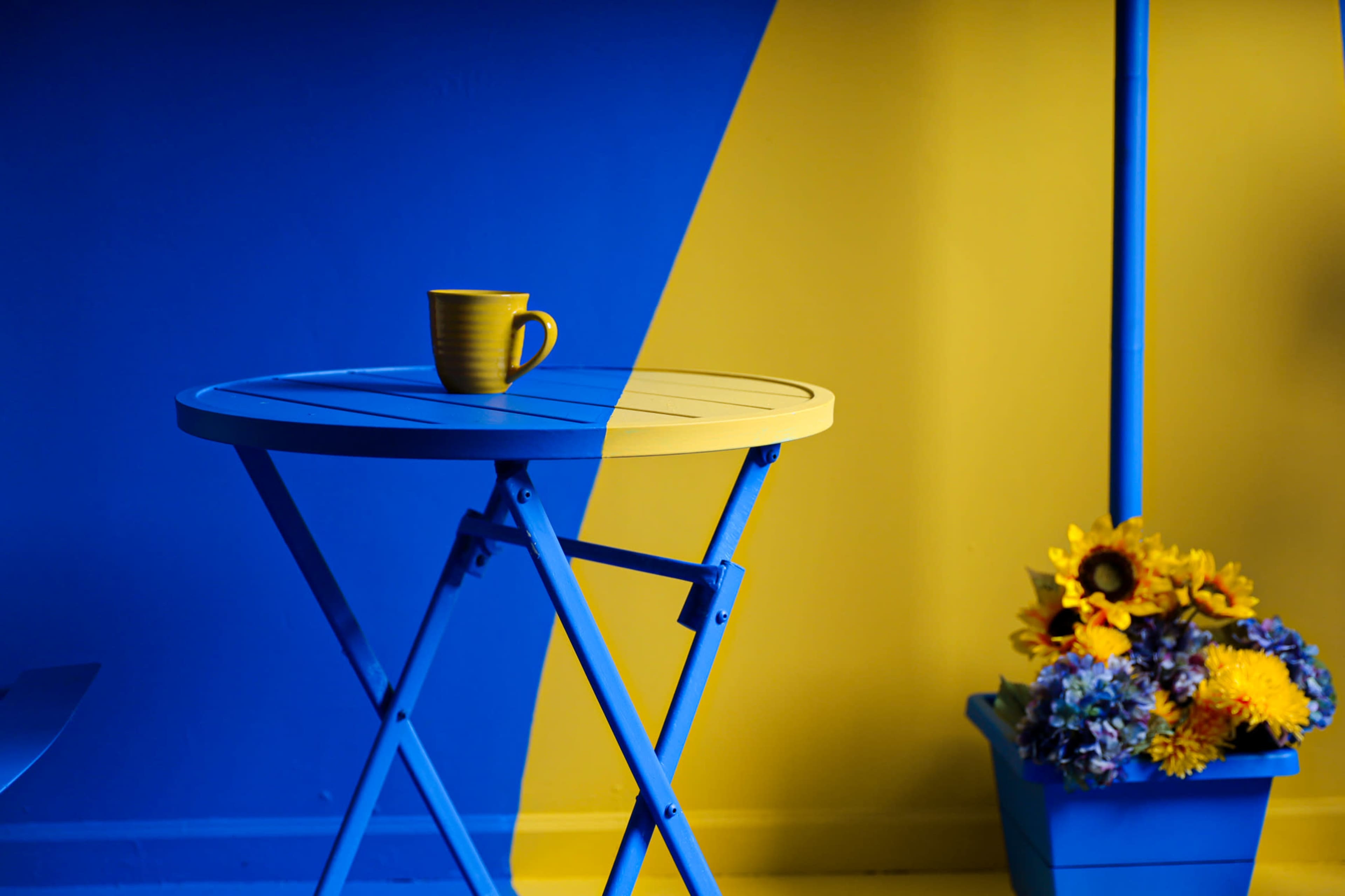 A blue and yellow room features a blue table with a yellow mug on top and a flower pot filled with bright blooms nearby.