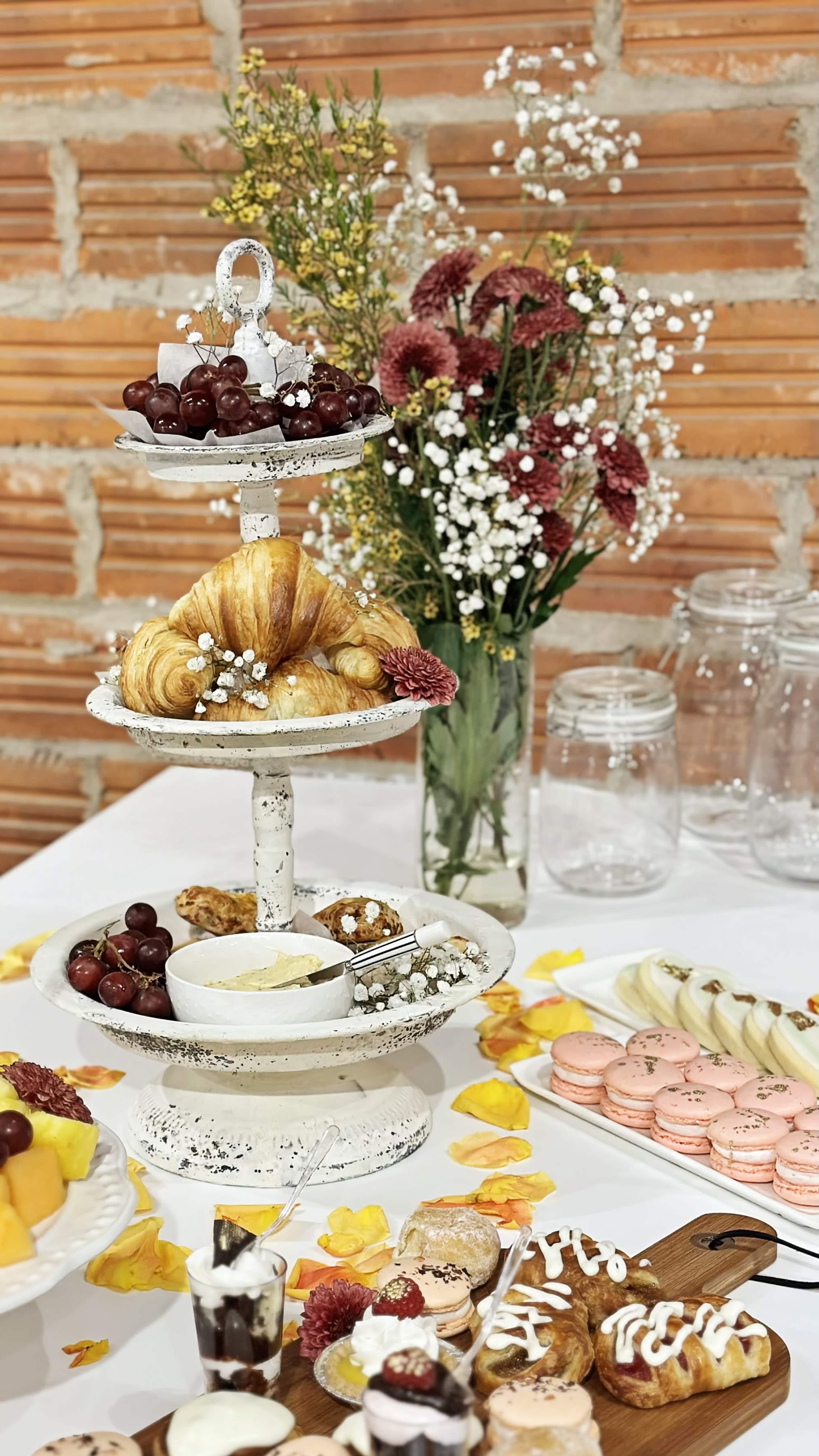 A three-tiered dessert stand displays croissants, grapes, and macarons, surrounded by a vase of flowers and jars on a table.