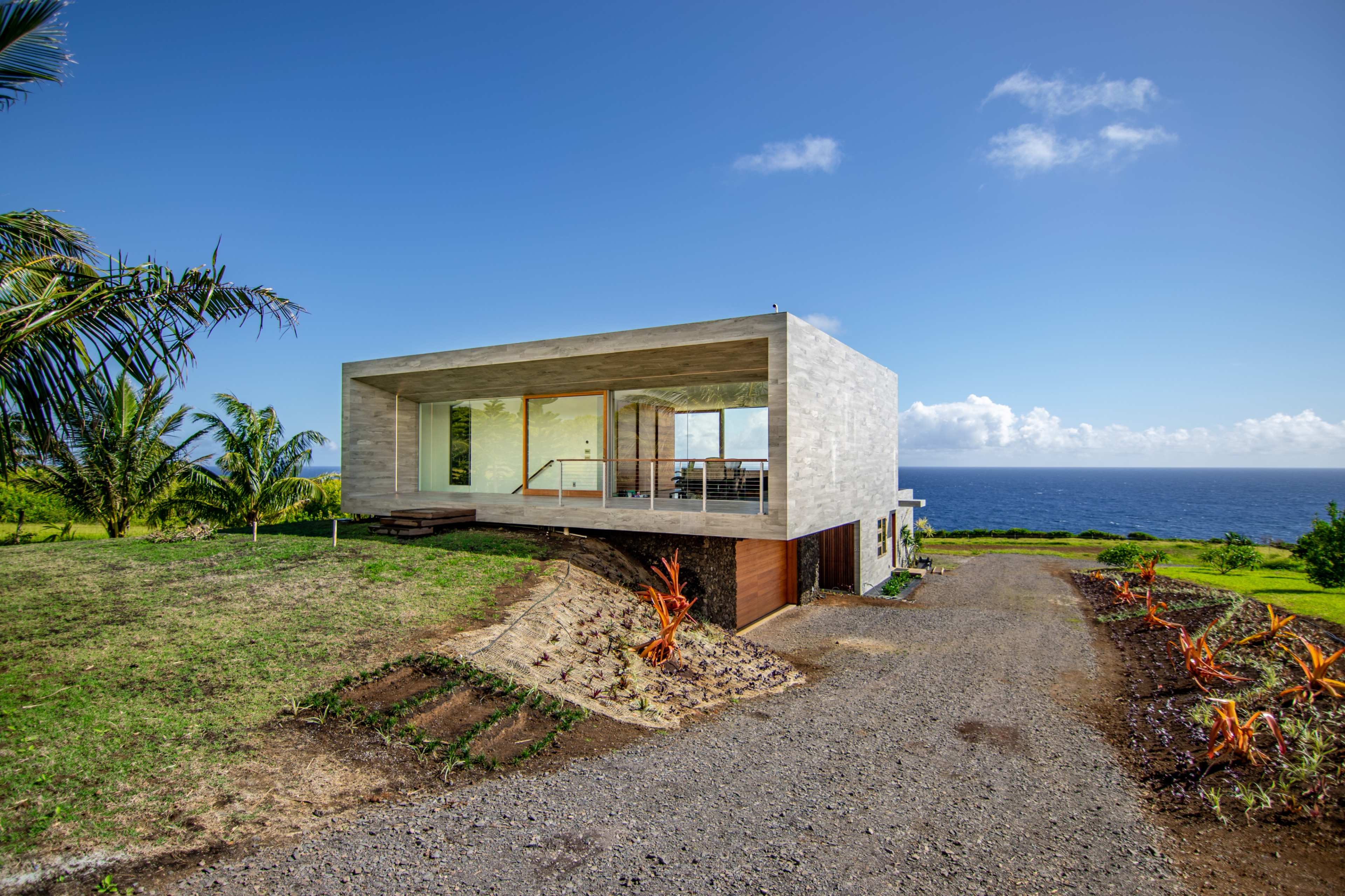 A modern concrete house with large glass windows is situated on a grassy hill overlooking the ocean.