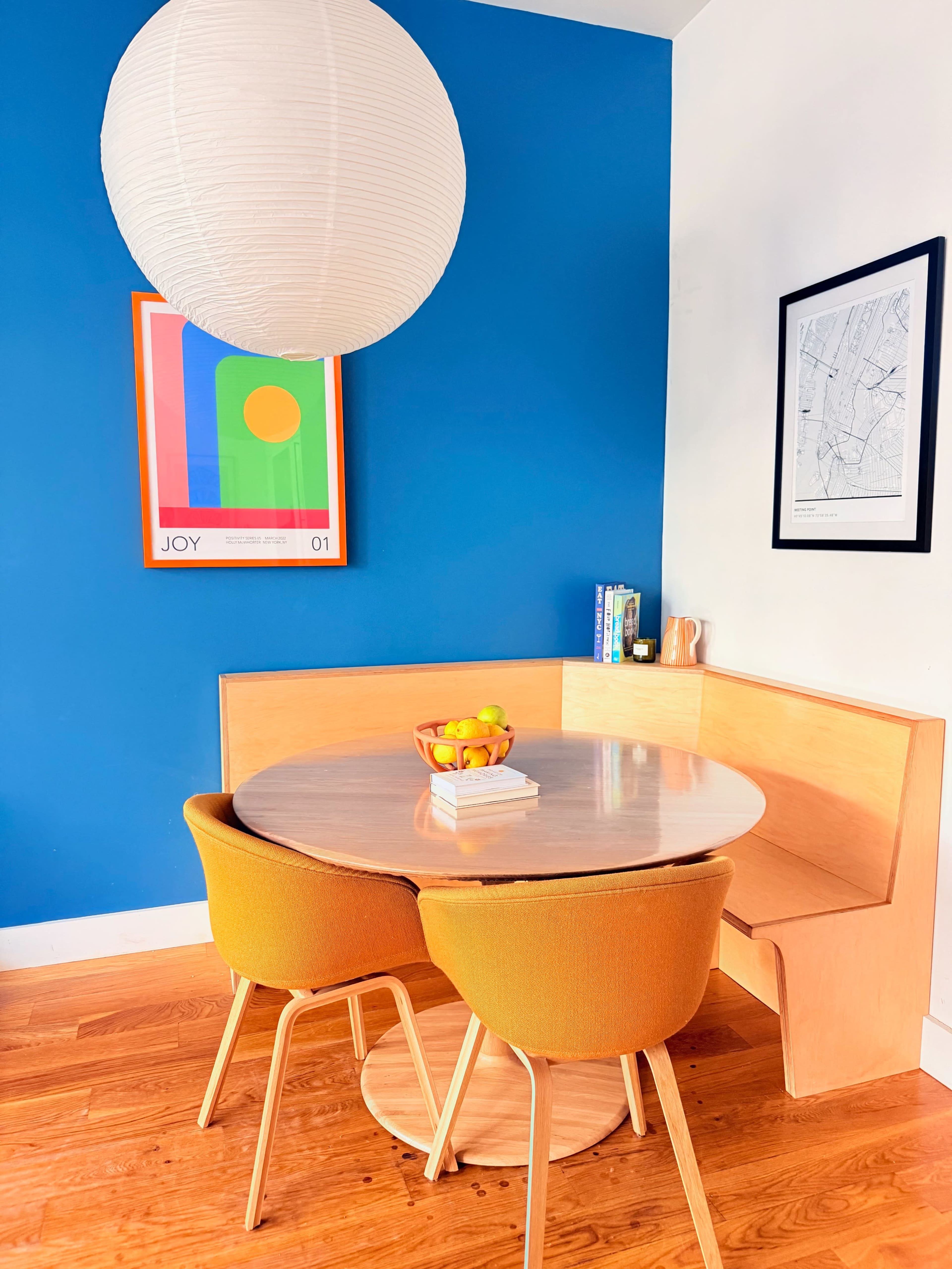 A round table with two orange chairs is positioned against a blue wall, complemented by a pendant light and framed artwork above.