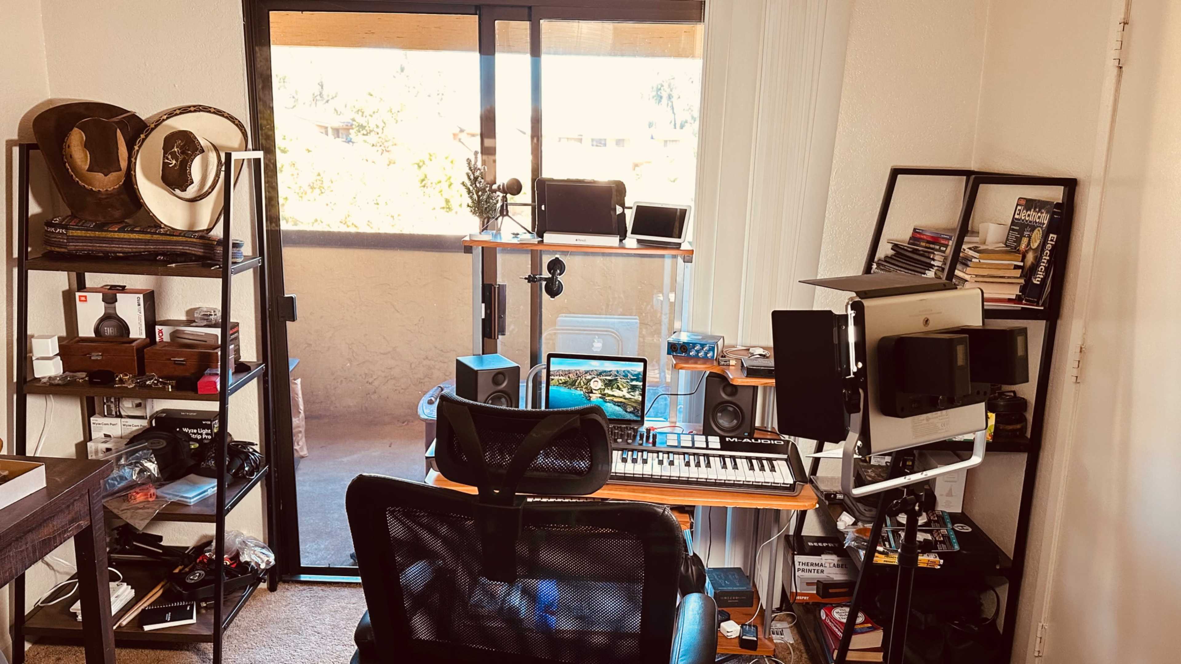 A workspace featuring a desk with a computer, audio equipment, and shelves filled with books and various items, alongside a large window leading to a balcony.