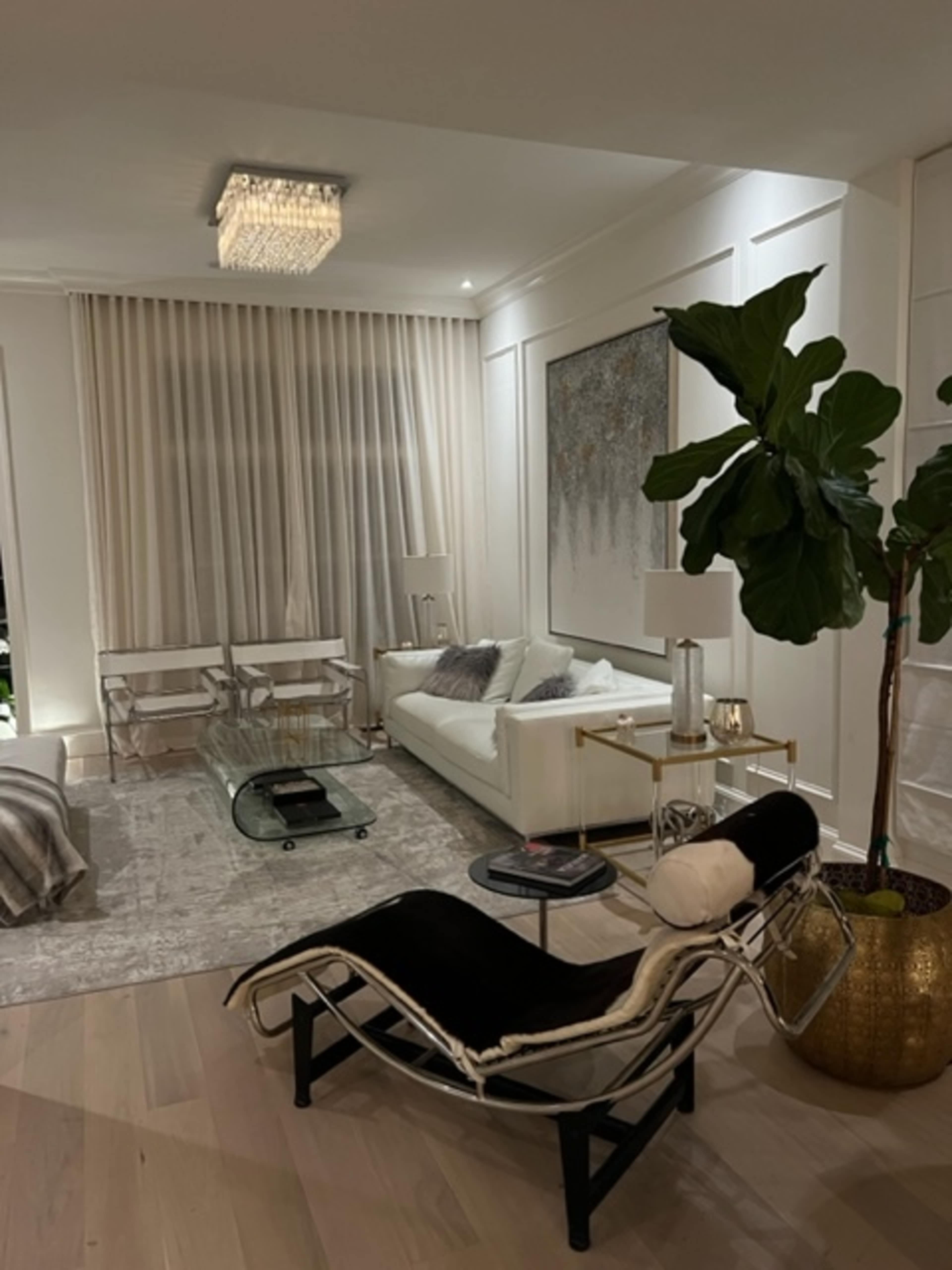 A modern living room with a white sofa, a glass coffee table, and a large indoor plant beside a window with sheer curtains.
