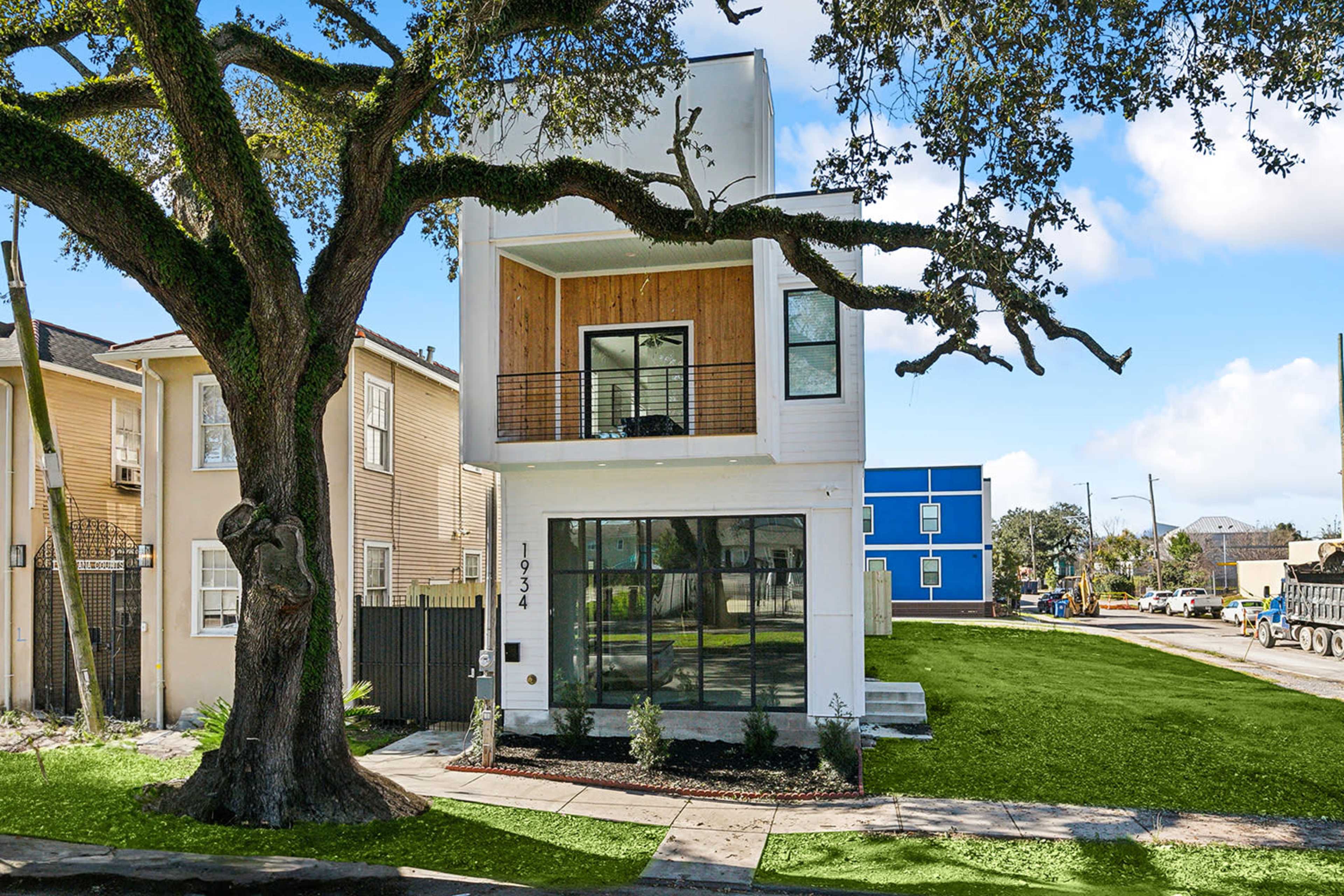A modern two-story house with large windows and a wooden balcony is situated next to a large tree, with a grassy yard and nearby residential buildings visible.