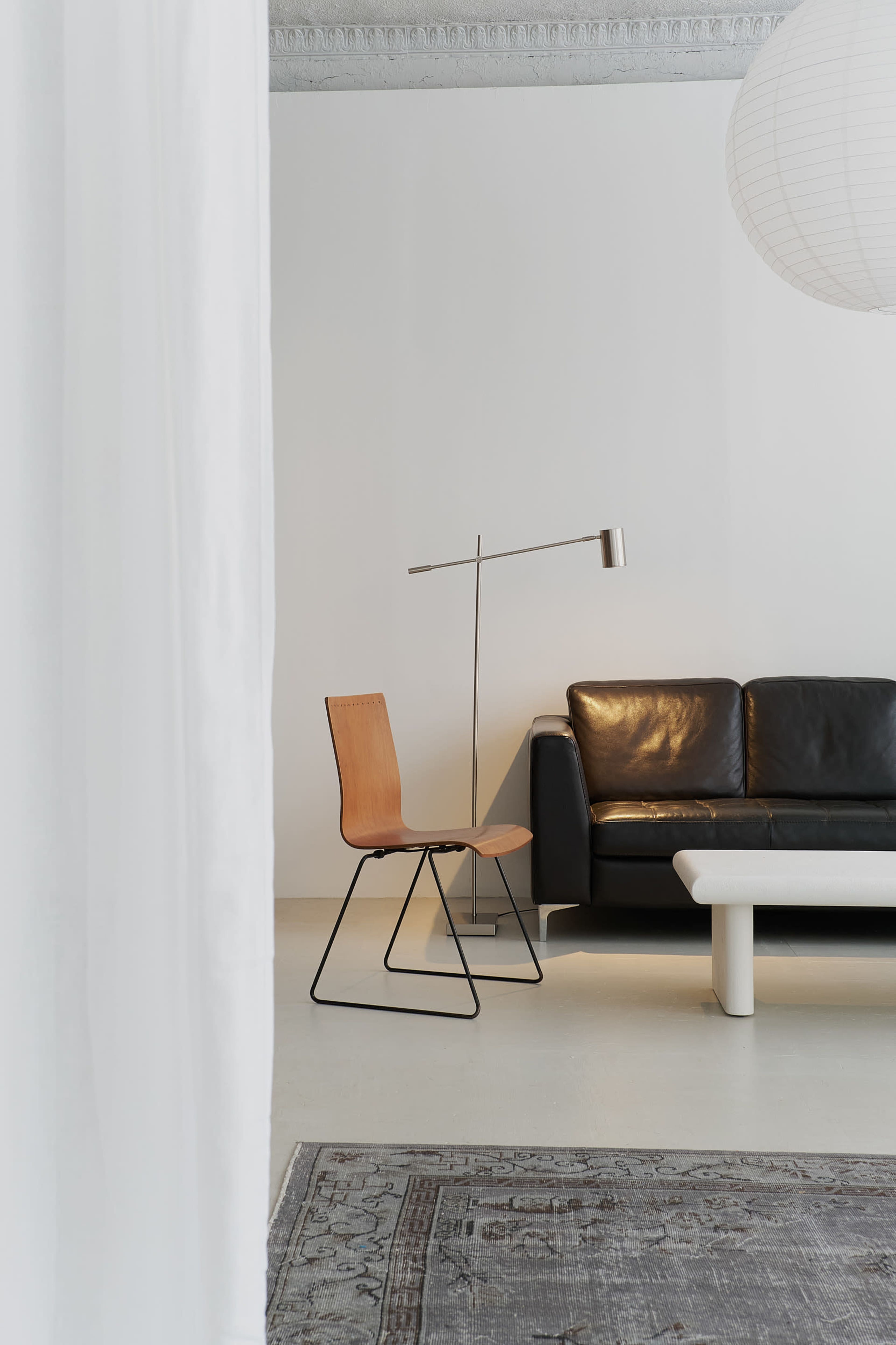 The image shows a minimalist living room featuring a black leather sofa, a light wooden chair on a metal frame, a white coffee table, and a floor lamp against a plain wall.