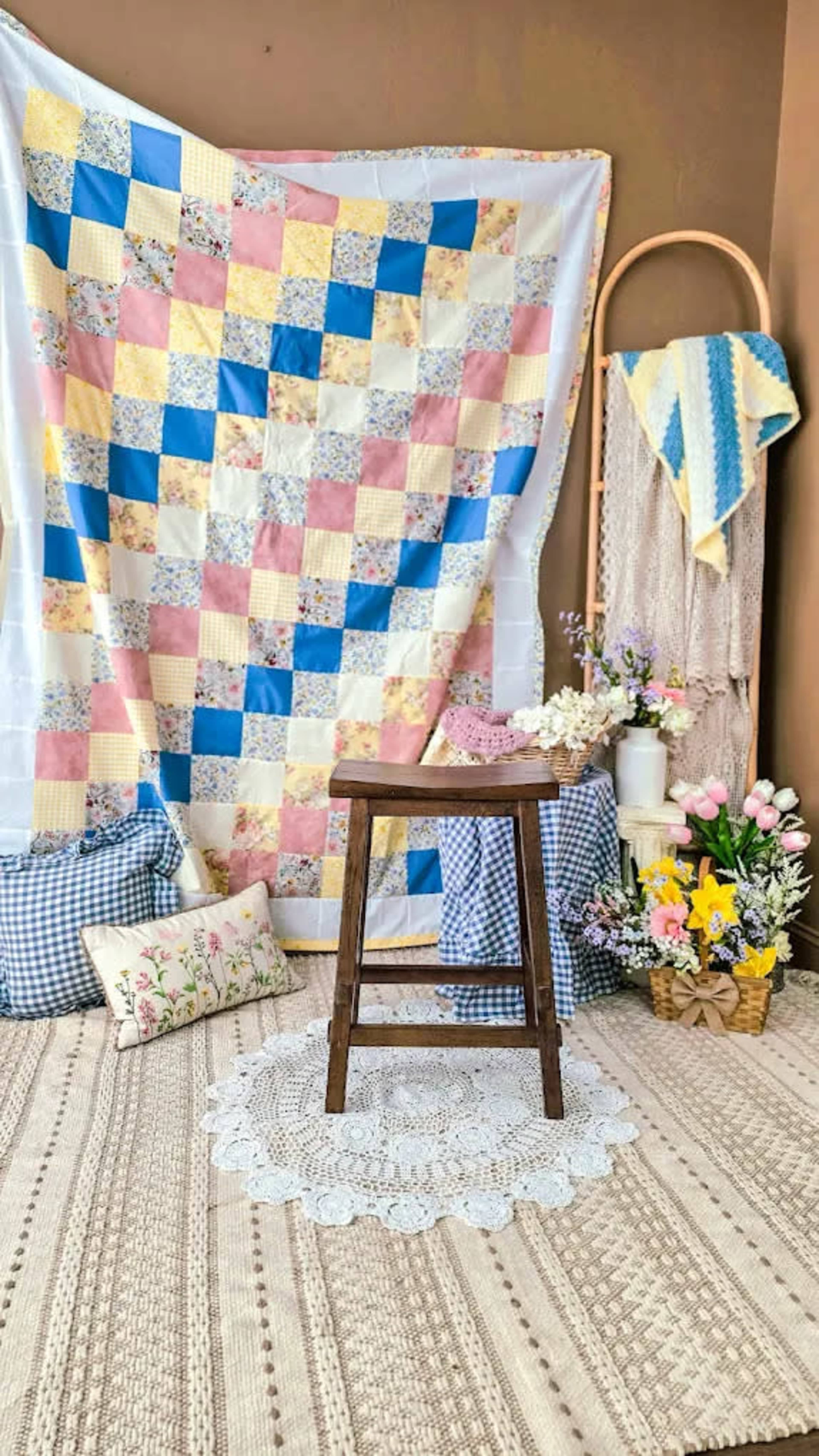 The scene features a quaint arrangement with a patchwork quilt backdrop, a wooden stool in the foreground, and a mix of decorative pillows and flowers on a light-colored rug.