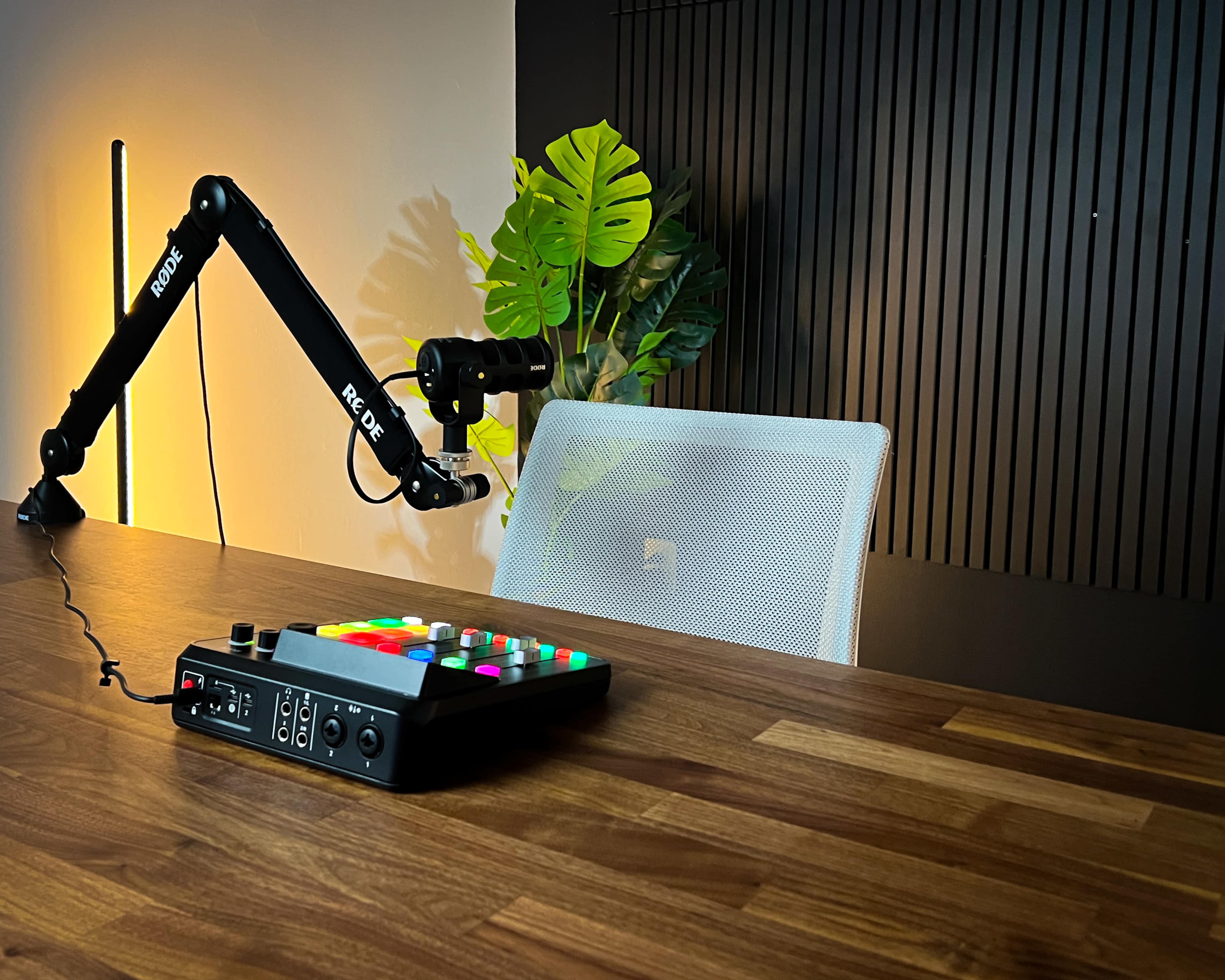 A black audio interface with colorful buttons is placed on a wooden desk next to a microphone arm, with a white chair and a plant in the background.