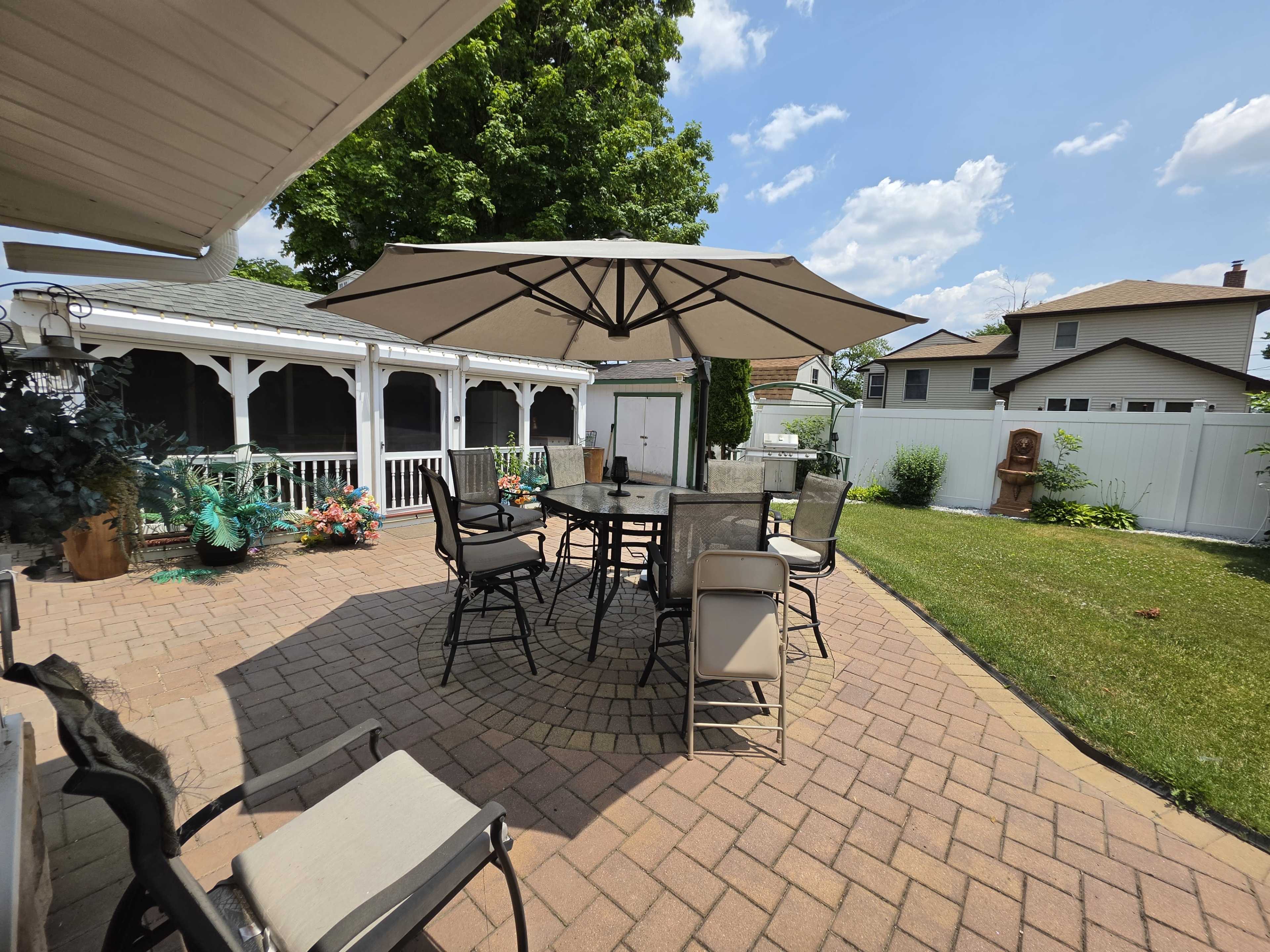 The image shows a well-designed patio area with a large umbrella over a dining table surrounded by chairs, set against a backdrop of green grass and houses.