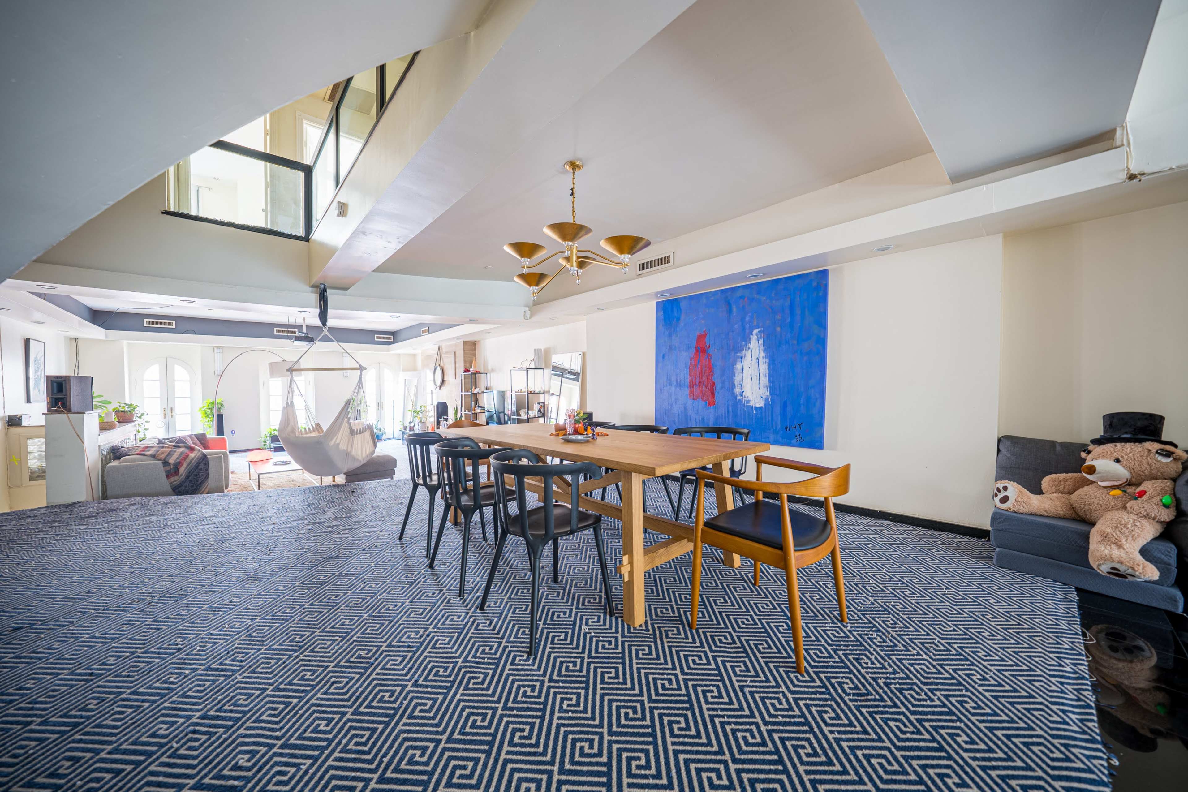 The image depicts a spacious indoor area with a large dining table surrounded by chairs, a hammock hanging nearby, and a prominent blue artwork on the wall.