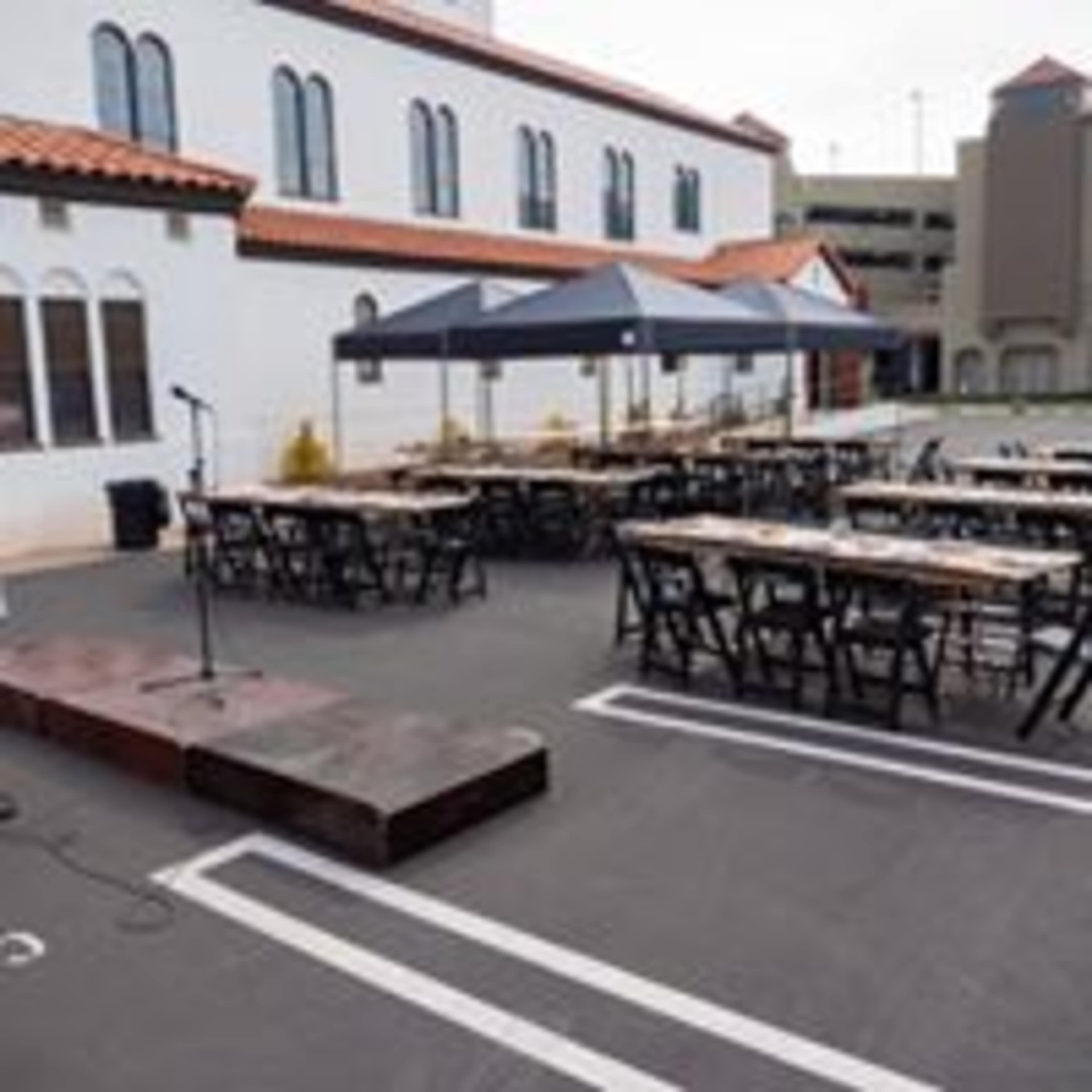 An outdoor event setup with rows of tables and chairs under two tents, accompanied by a microphone stand on a small stage, against a backdrop of a building and a parking structure.