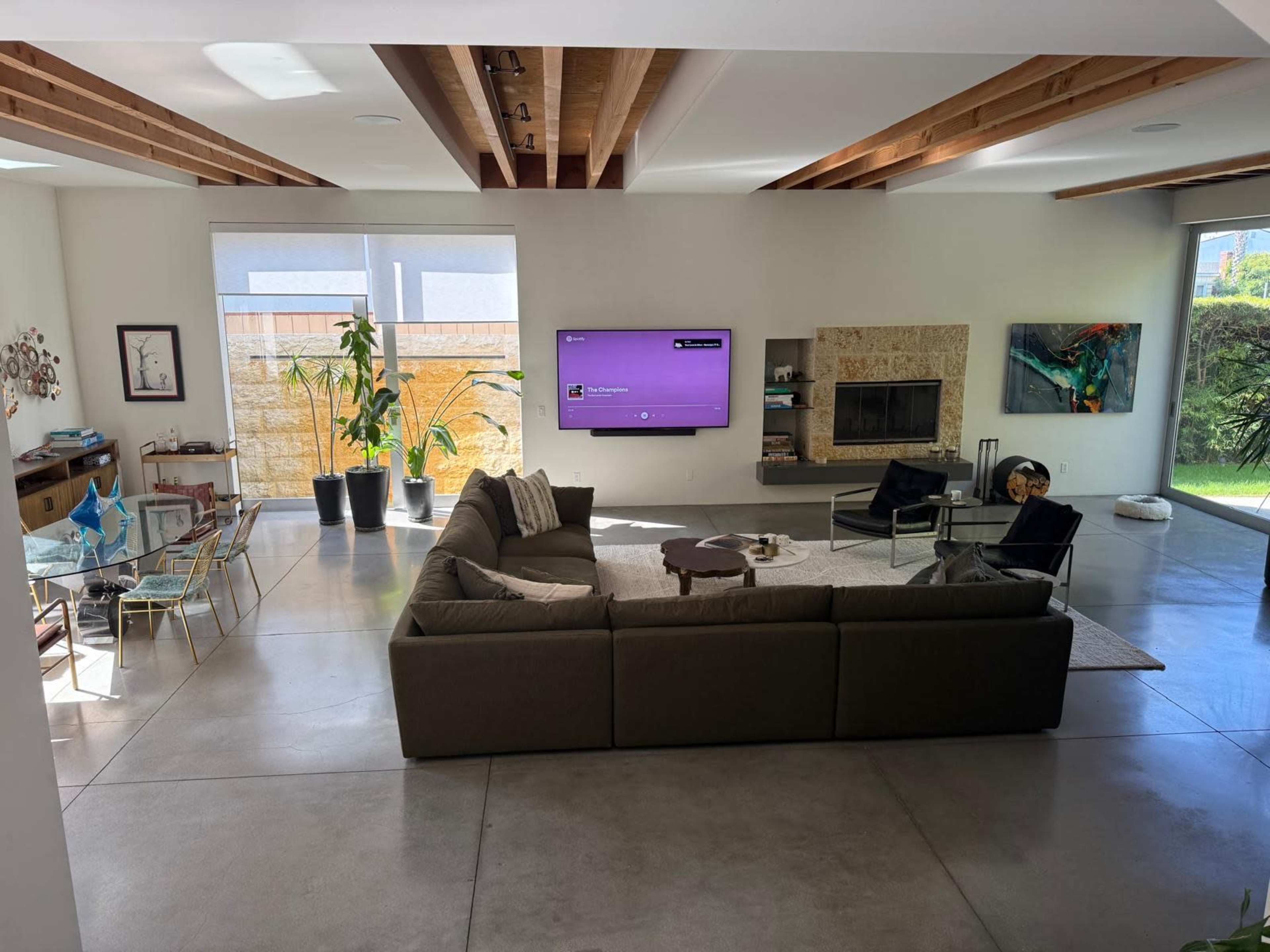 The image shows a modern living room with a large sectional sofa, a coffee table, a TV mounted on the wall, and a fireplace, surrounded by plants and contemporary decor.