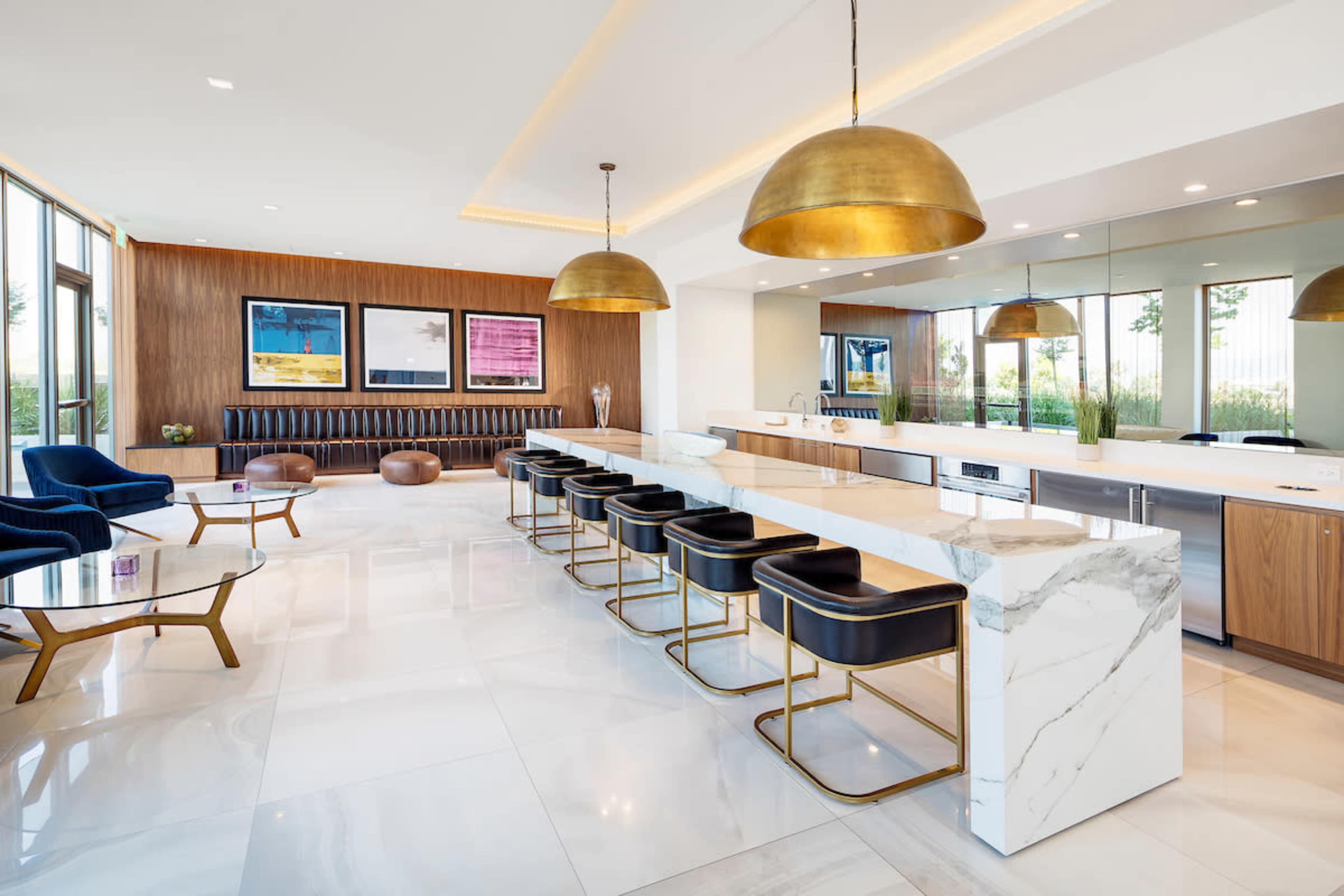A modern kitchen area features a marble countertop bar with gold pendant lights and black stools, surrounded by sleek furniture and large windows.
