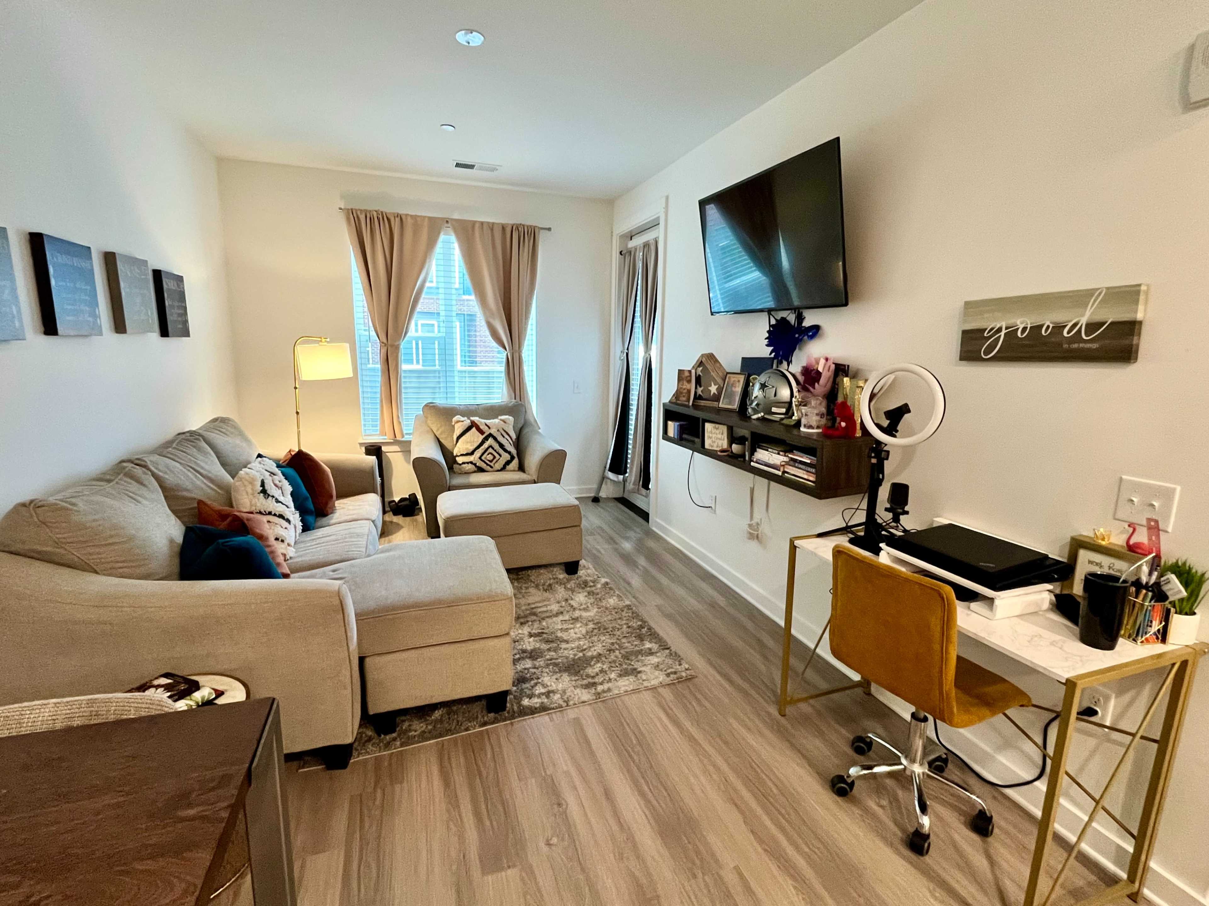 The image shows a cozy living room with a beige sectional sofa, two chairs, a wall-mounted TV, and a desk with a chair, all arranged in a well-lit space with curtains and decorative elements.