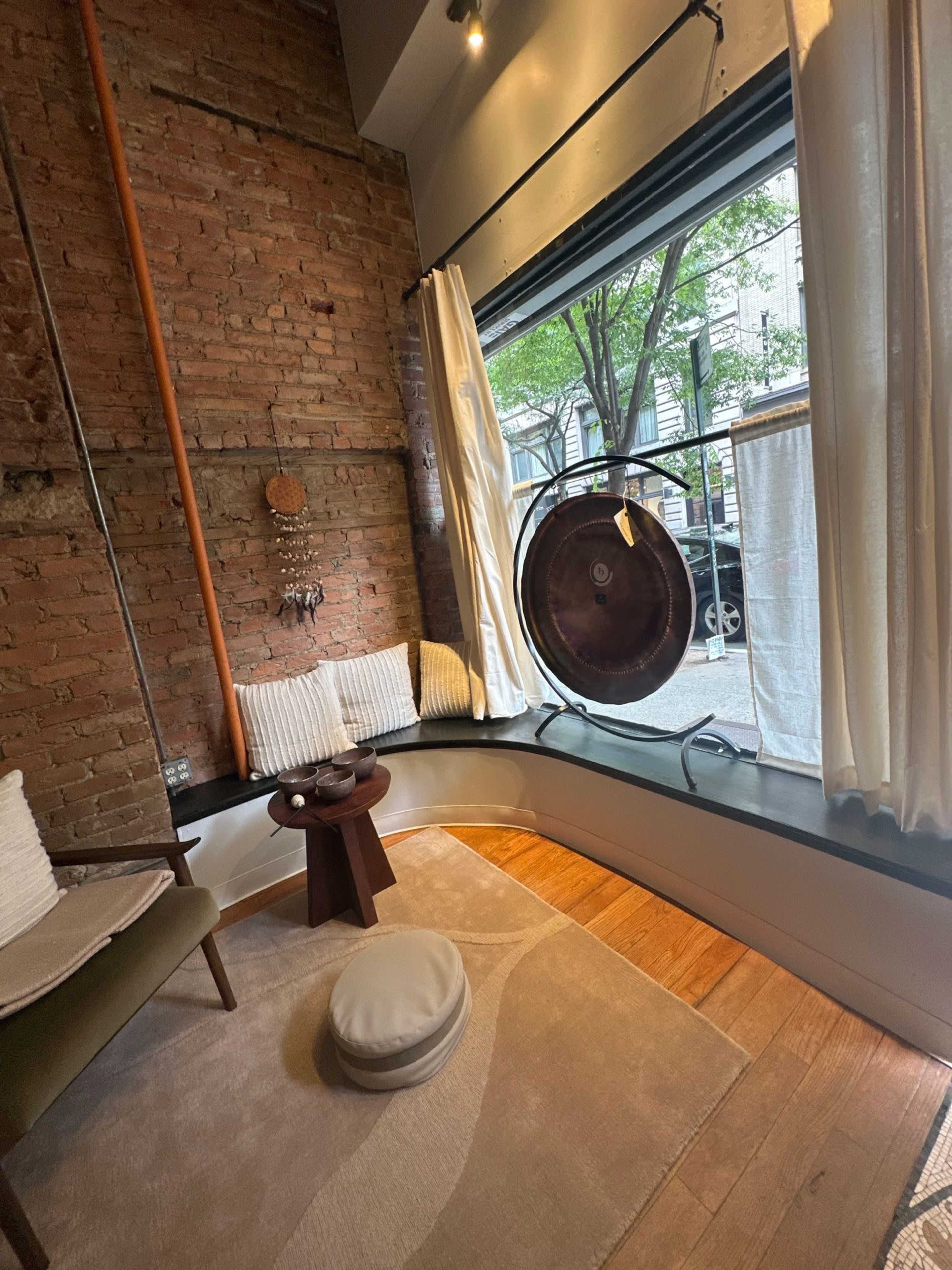 A cozy corner of a room features a circular window with a view of the street, a small wooden table, and a plush seating area adorned with cushions and a rug.