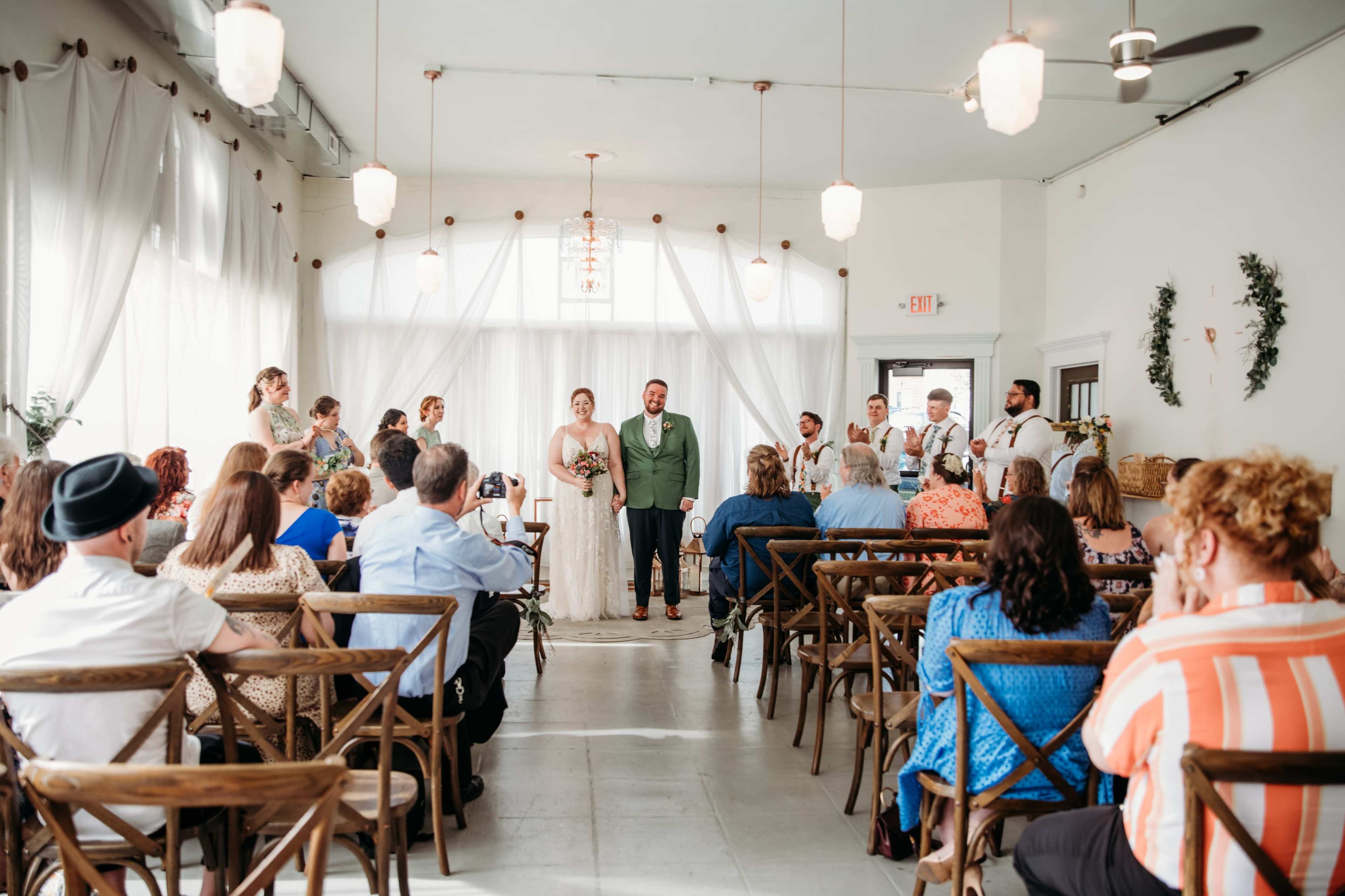 A wedding ceremony is taking place in a well-lit venue, with guests seated on wooden chairs and the bridal party standing at the front.