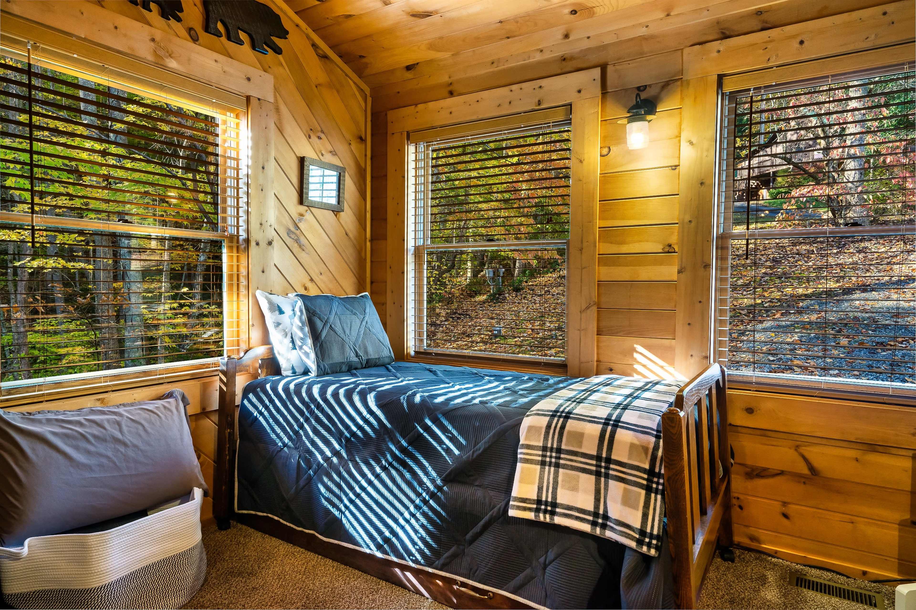 The image shows a small, cozy bedroom with wooden walls, large windows, and a single bed covered with a dark blanket and a plaid throw.