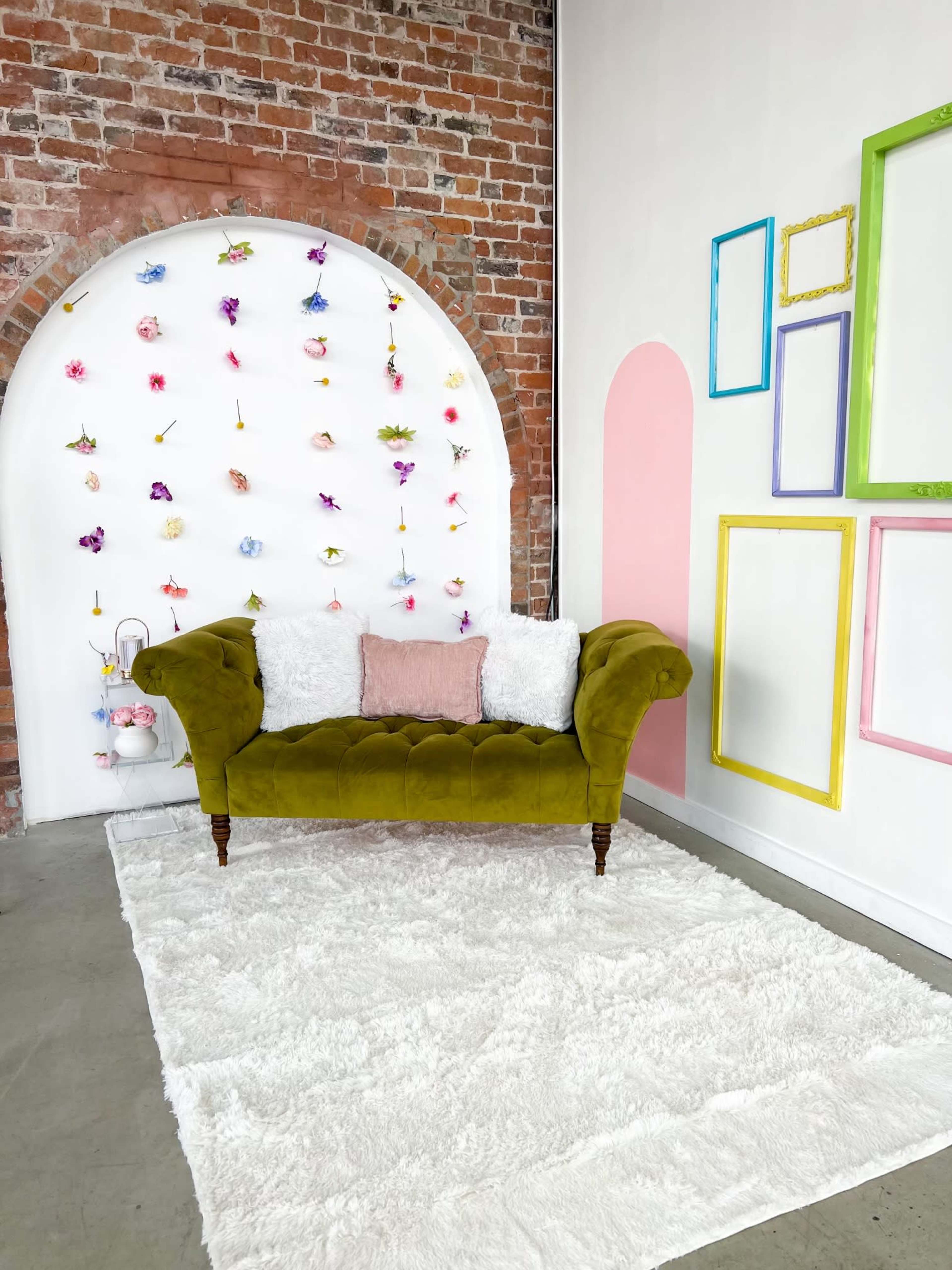 A green velvet sofa with white cushions placed on a fluffy rug in front of a wall decorated with colorful flowers and framed outlines.