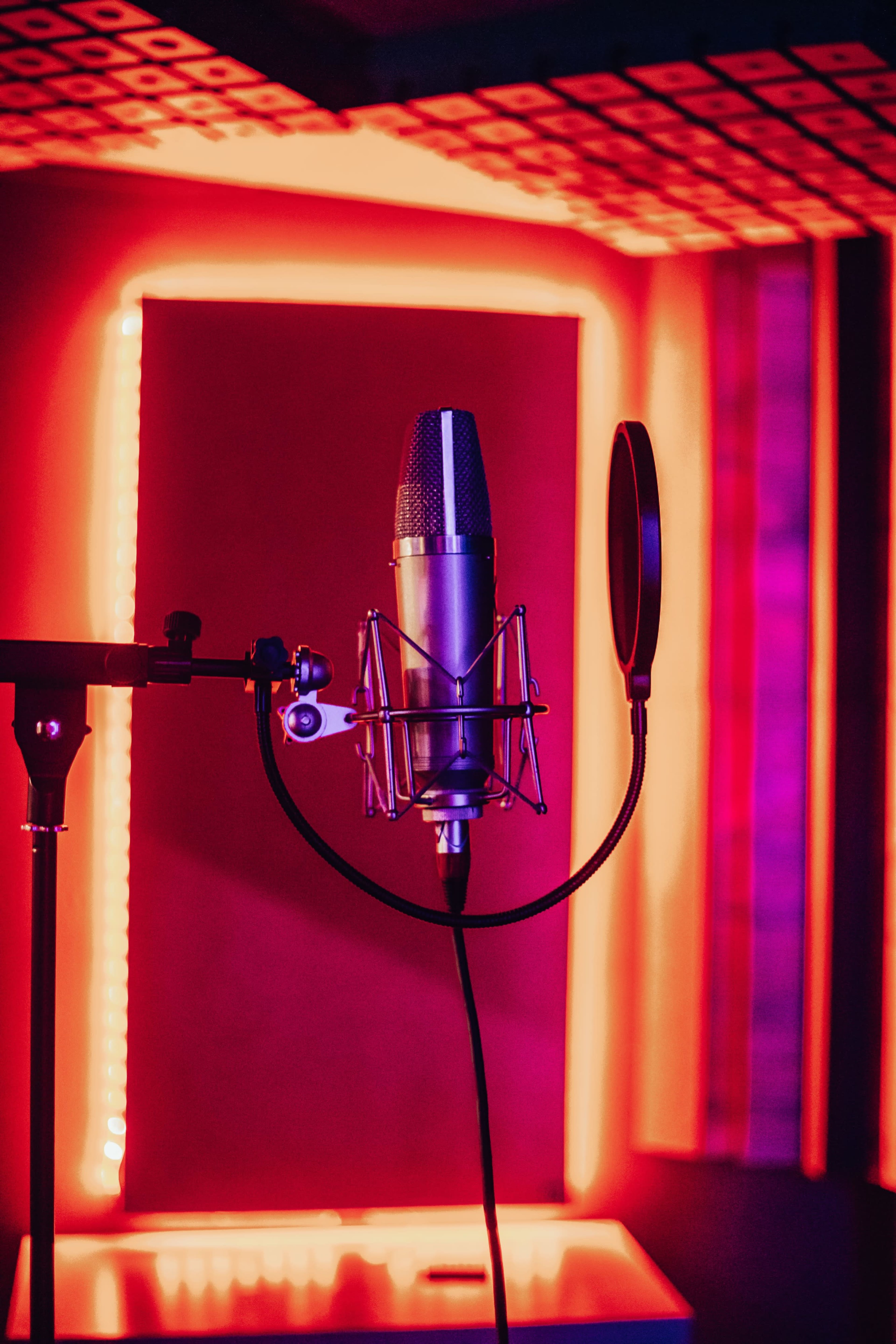 A microphone is mounted on a stand in a sound studio illuminated by red and purple lighting.
