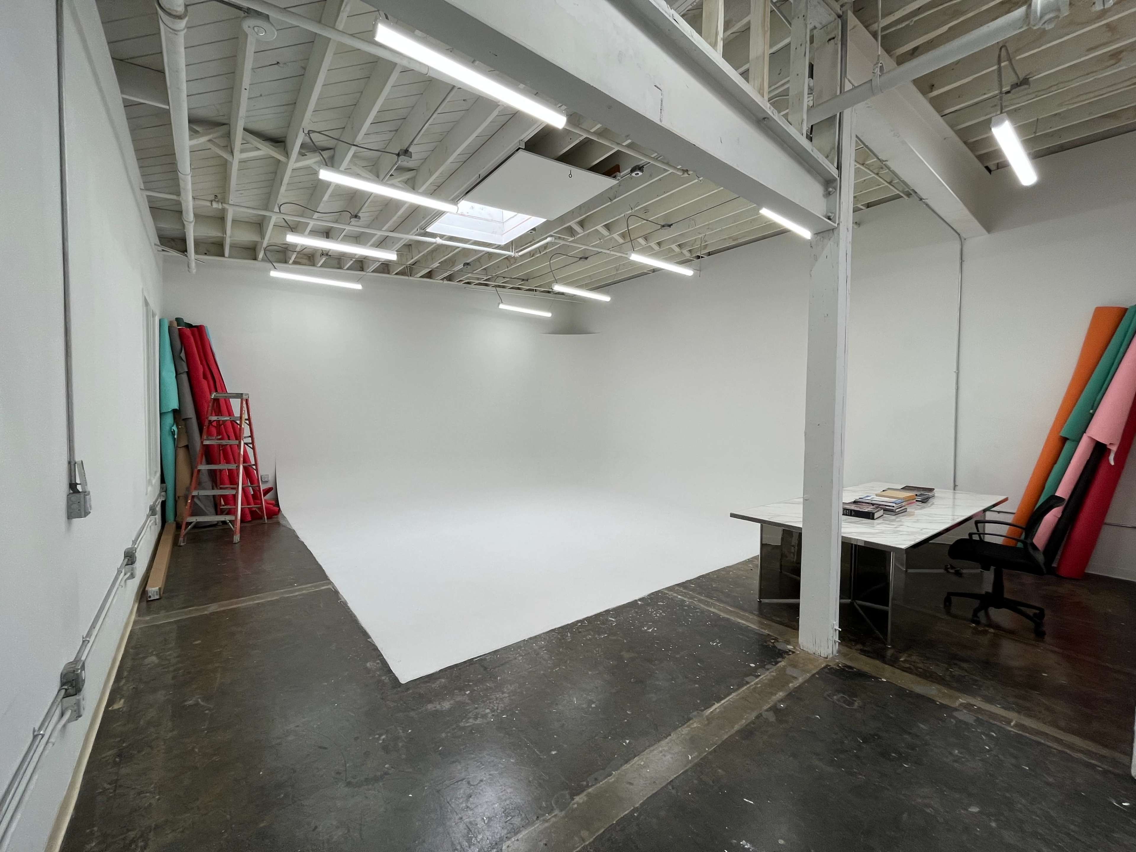 The image shows a spacious, unfinished studio with a white backdrop, a desk with a chair, and colorful rolls of paper along the wall.