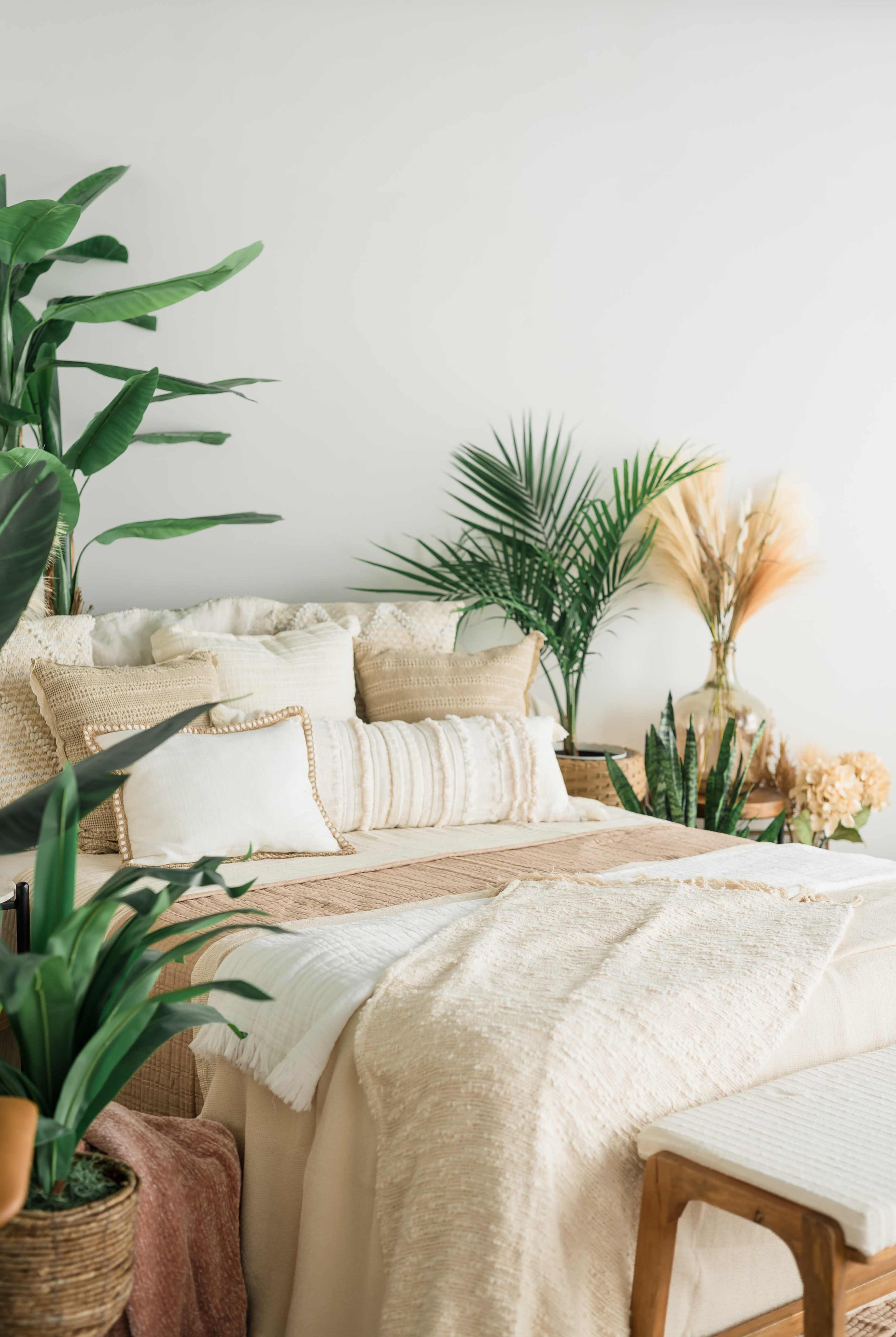 A well-decorated bedroom features a bed with neutral-toned pillows and throws, surrounded by various indoor plants against a light-colored wall.