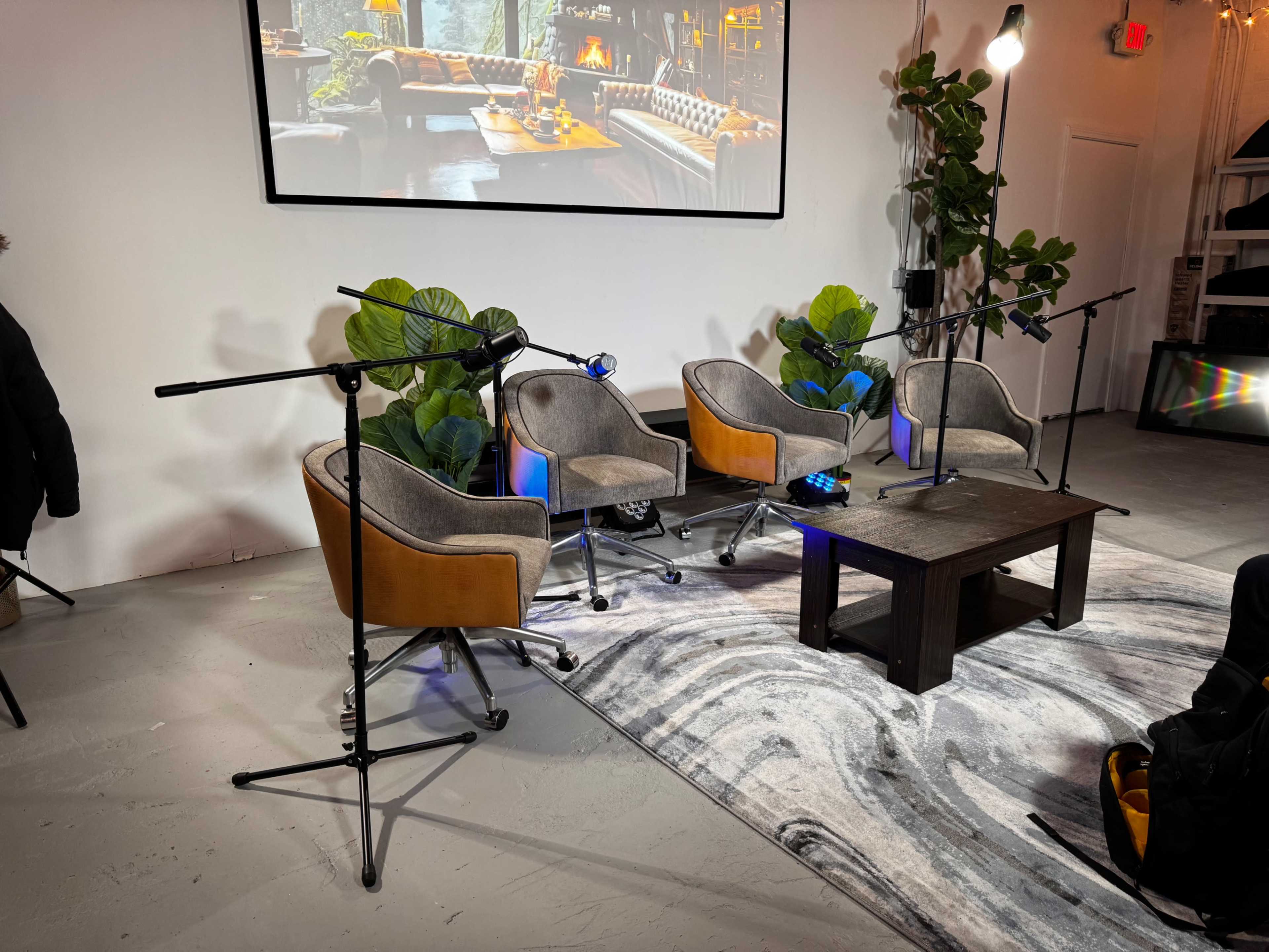 A cozy event space with four upholstered chairs arranged around a low wooden table, equipped with microphones and surrounded by large green plants.