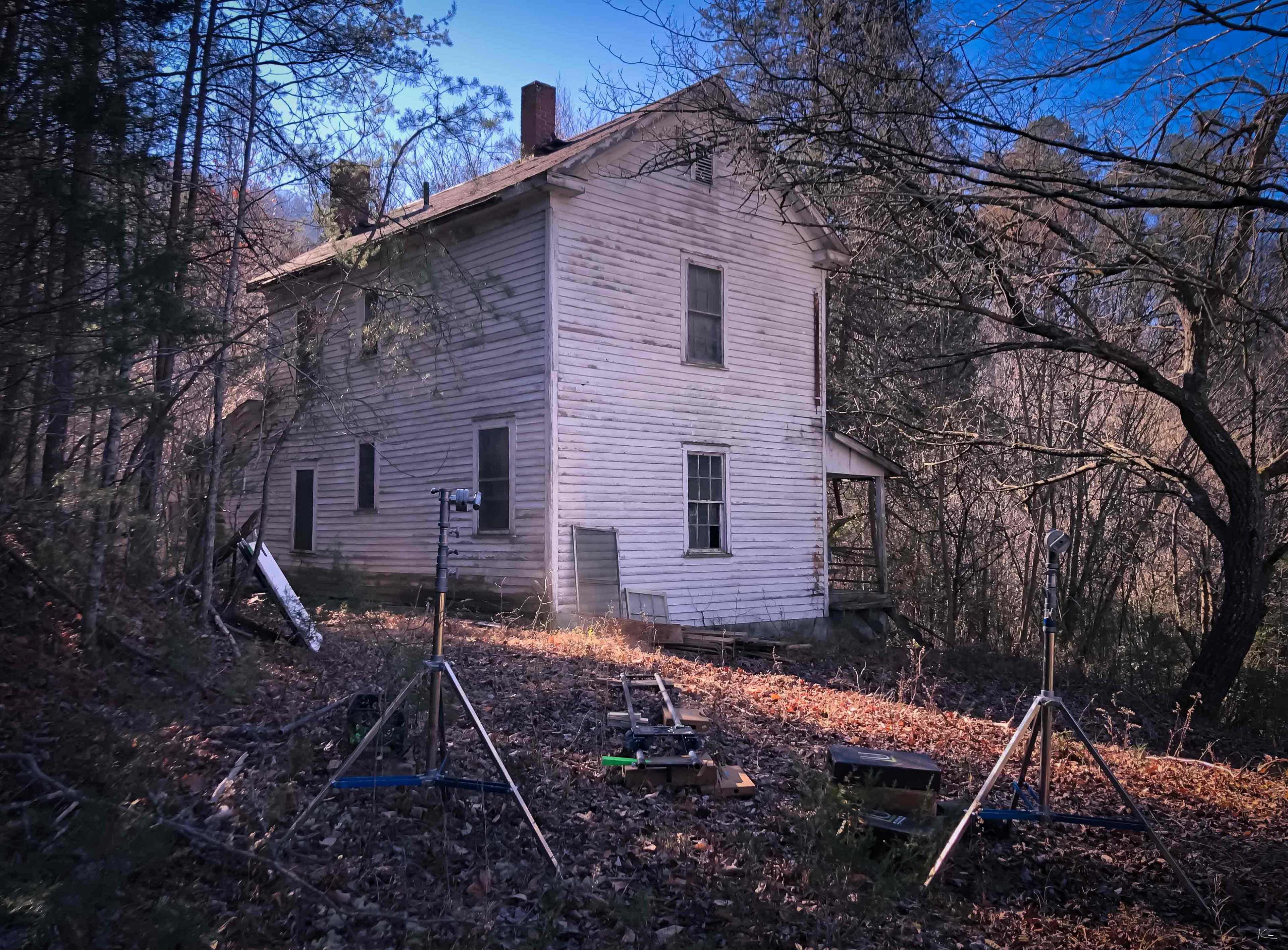 The image shows a weathered, two-story white house surrounded by trees and debris, with equipment set up in the foreground.