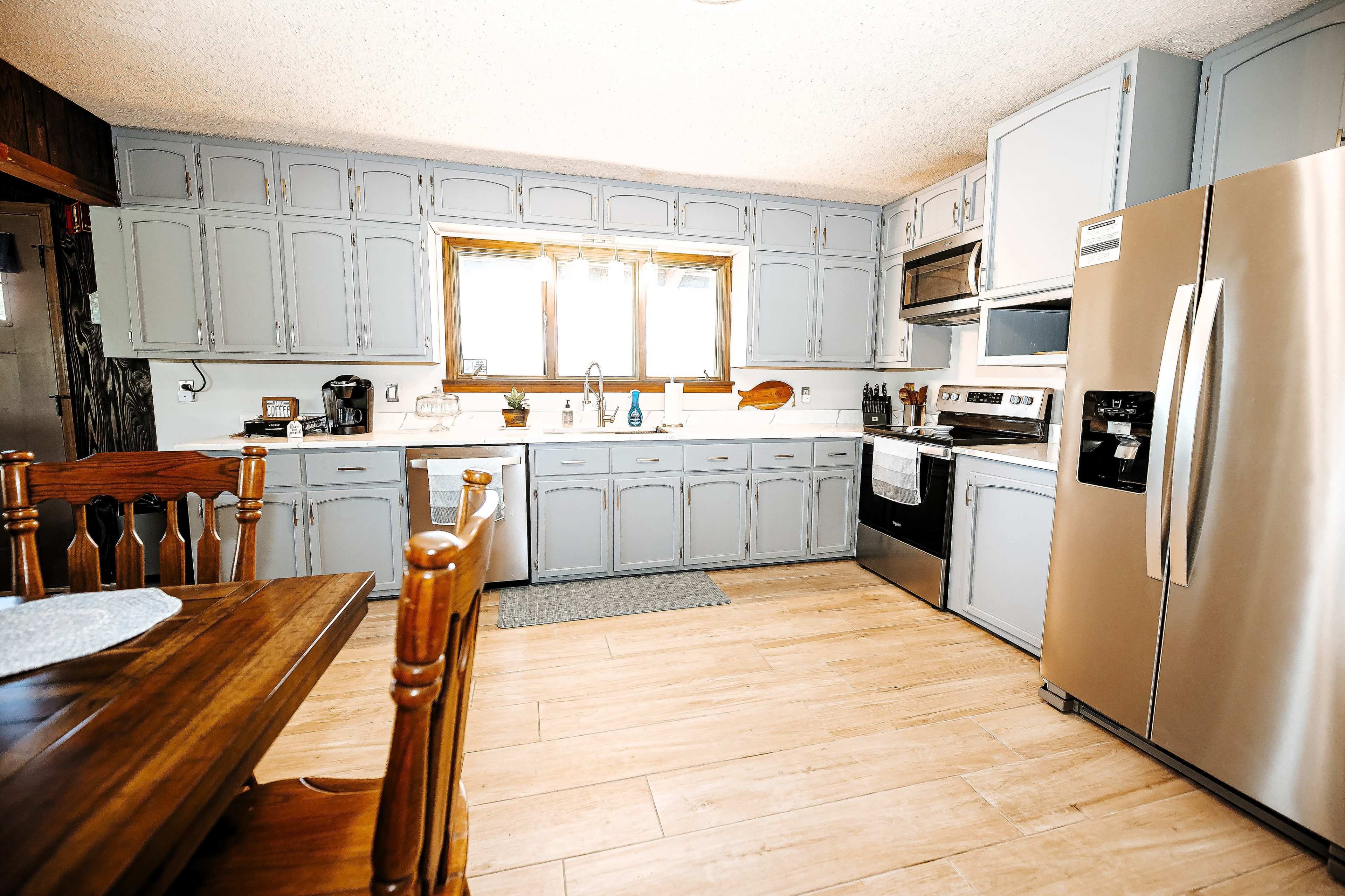 The kitchen features light gray cabinets, stainless steel appliances, and a wooden dining table set against a backdrop of tiled flooring.