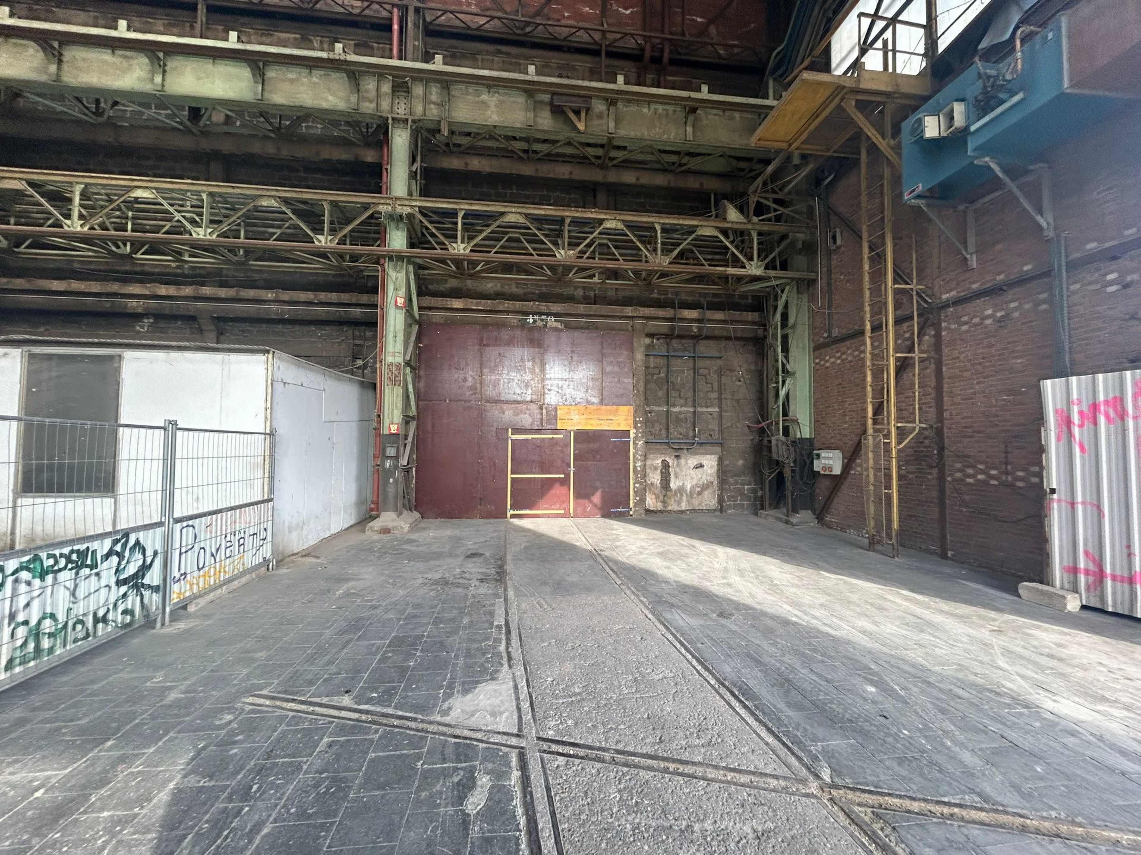 An empty industrial space with a large metal door and exposed brick walls, illuminated by sunlight.