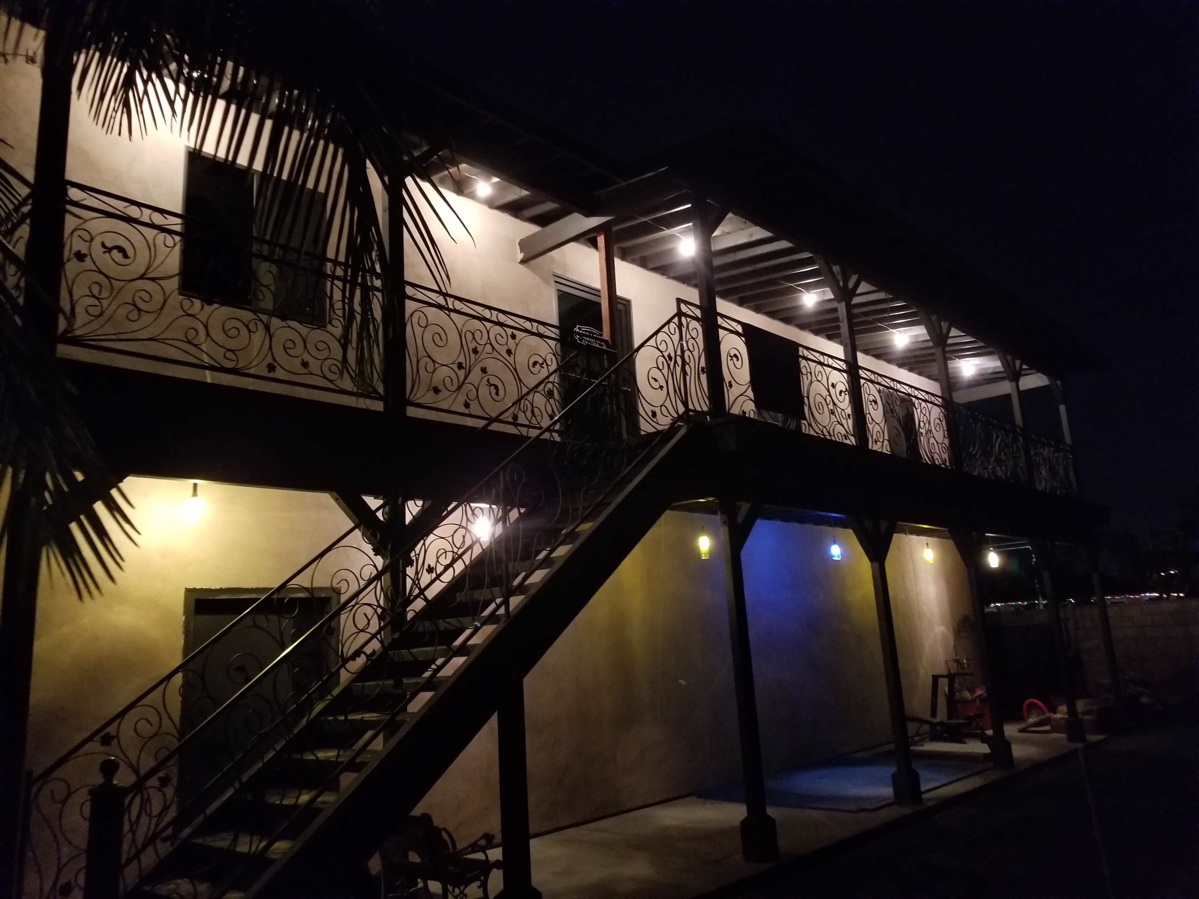 A two-story building with a staircase and decorative balcony lighting illuminates the exterior at night.
