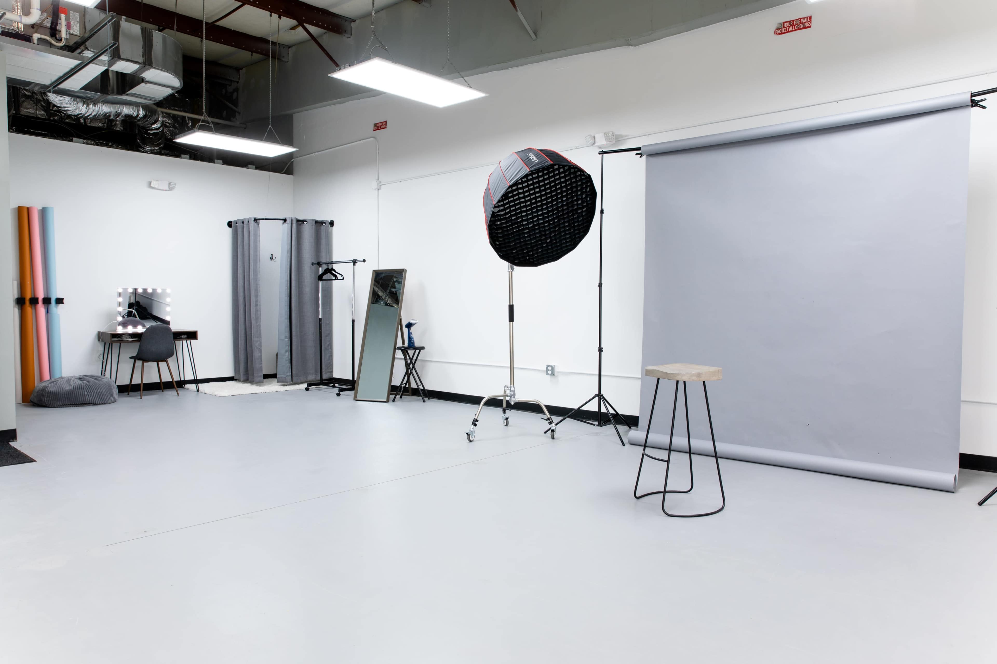 A photography studio with a gray backdrop, lighting equipment, a mirror, a chair, and a small table in a spacious, well-lit setting.